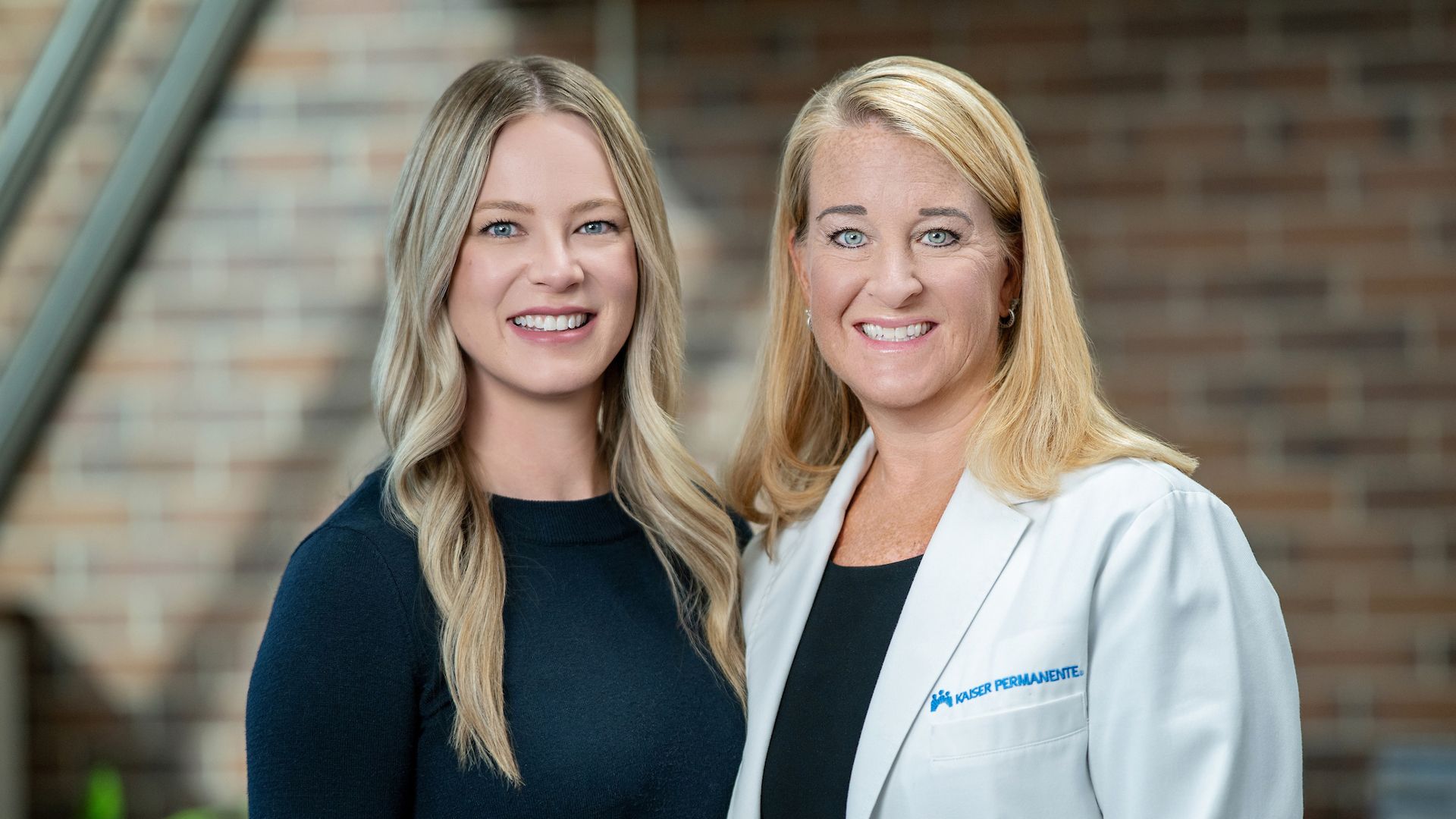 Two smiling blonde women pose indoors; left in a dark navy top, right in a white lab coat with the Kaiser Permanente logo, brick wall background.