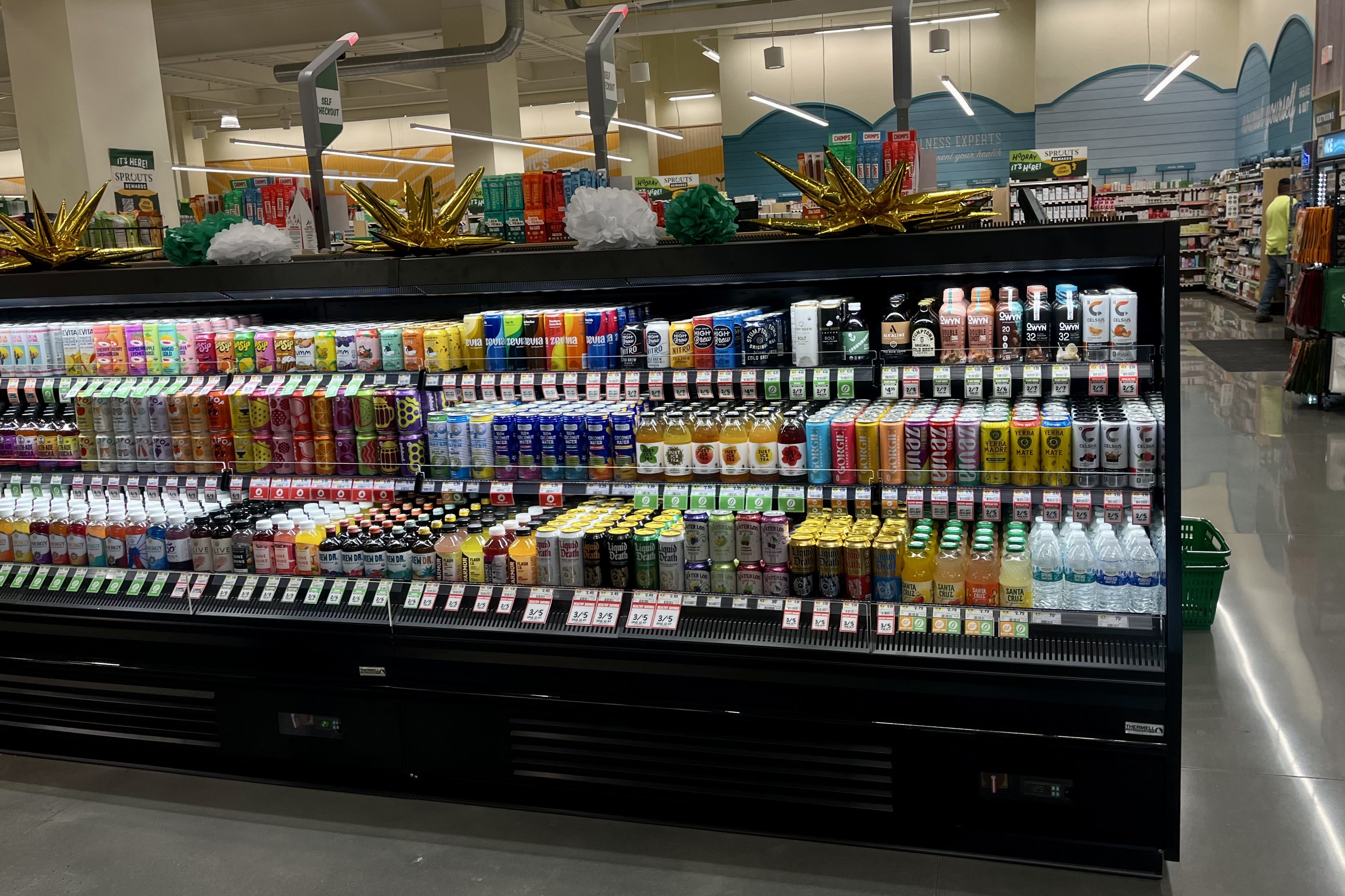Grocery store refrigerated shelf with various colorful canned and bottled drinks including soda, kombucha, and flavored water, decorated with gold and tissue paper ornaments on top.