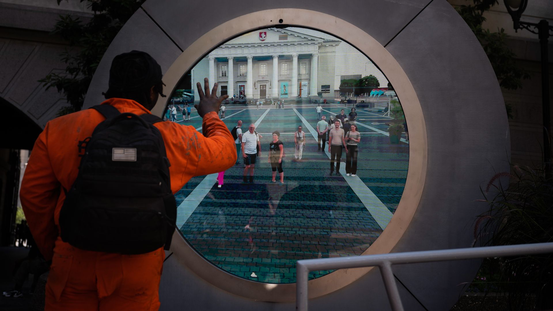 Back view of a person in an orange jacket and backpack reaching toward a circular display. The screen shows a plaza with a grand neoclassical building, columns, and crowds.