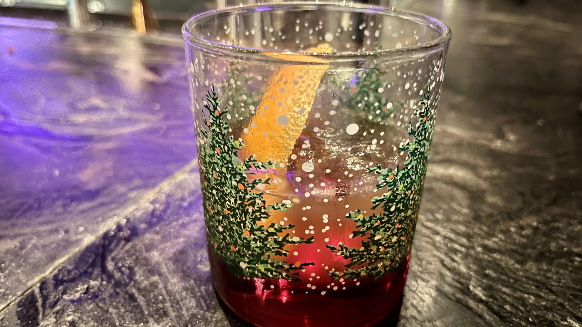 A red-colored cocktail and an orange slice in a tumbler with Christmas trees on it.