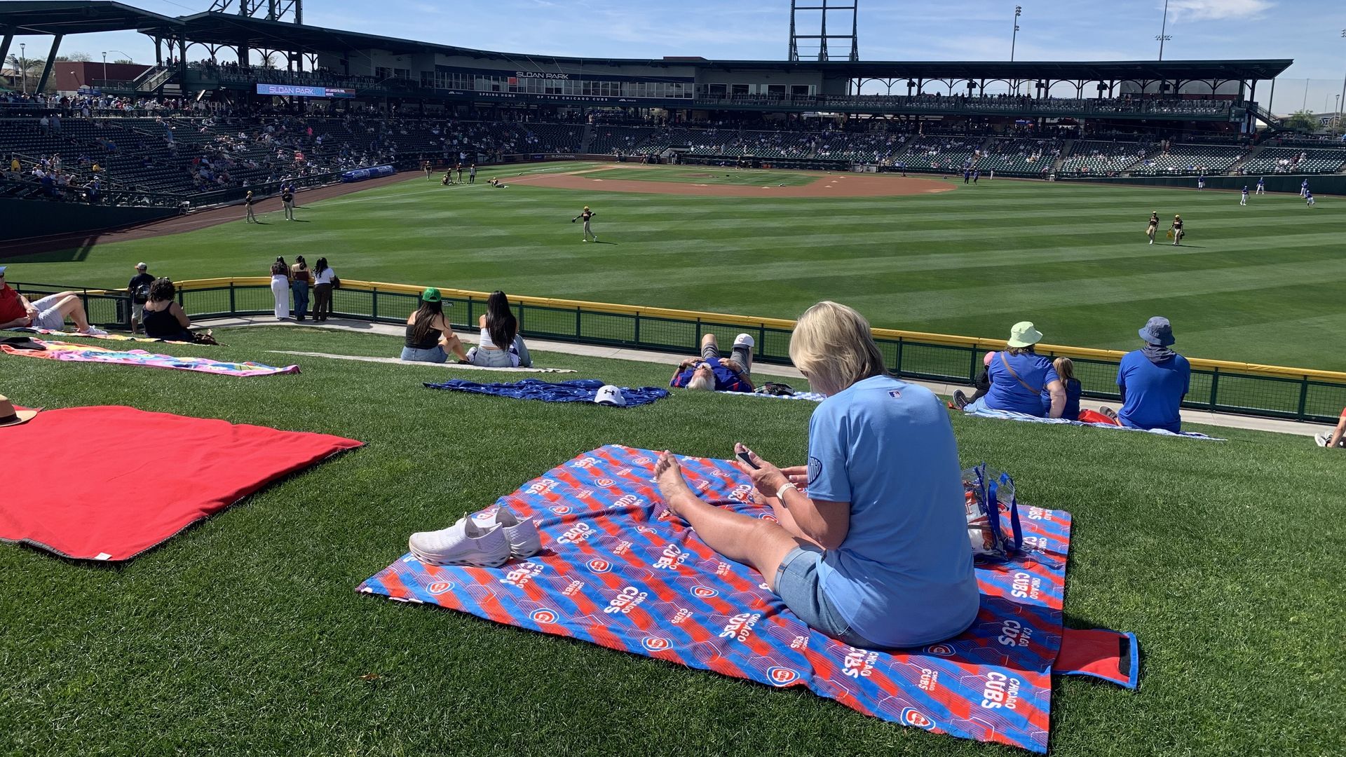 People sit on a grassy hill covered with Chicago Cubs blankets, watching a baseball game at Sloan Park under a sunny blue sky with scattered white clouds.