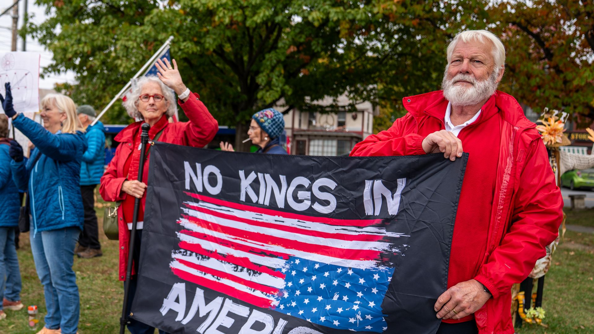 A man and a woman in red jackets hold a sign that reads "NO KINGS IN AMERICA" with an upside-down American flag.