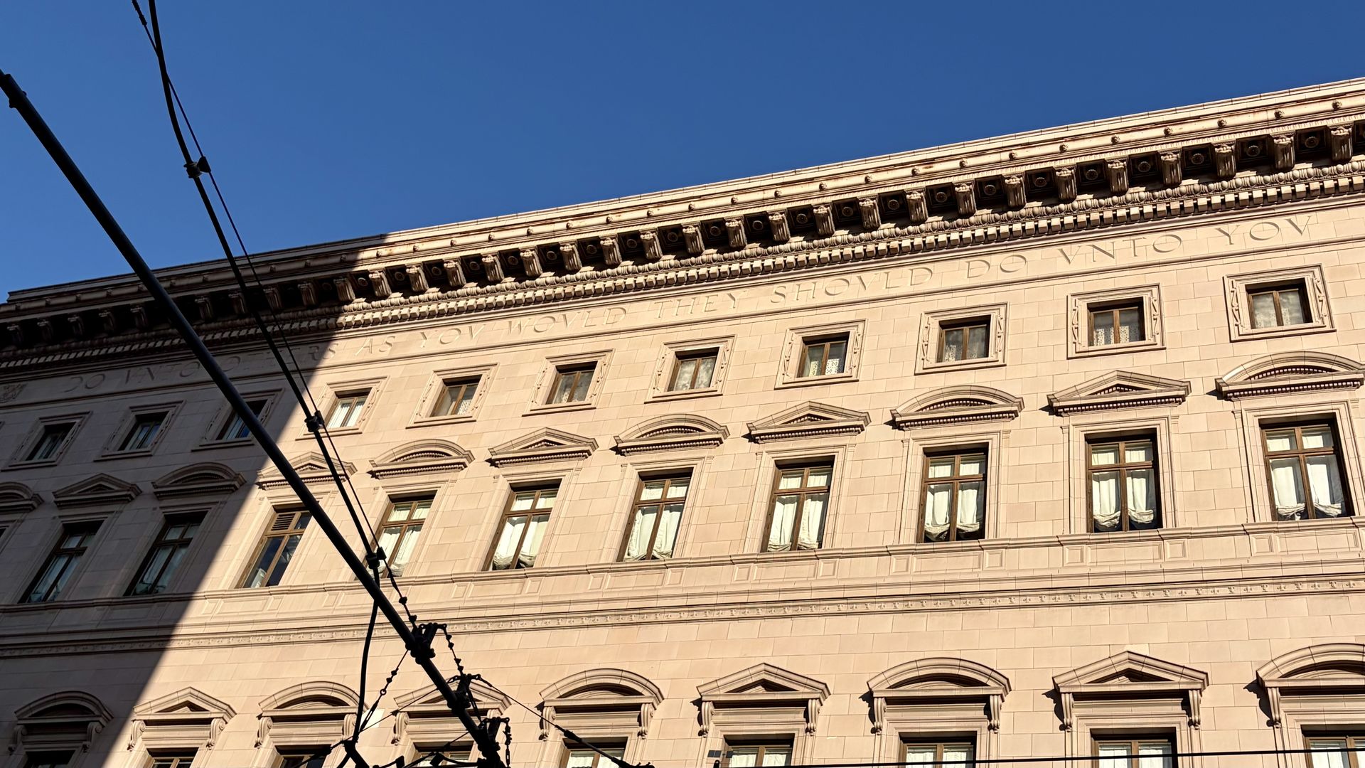 A sunlit, historic beige stone building with ornate cornice and large windows, framed by a clear blue sky; overhead tram cables cross the foreground.