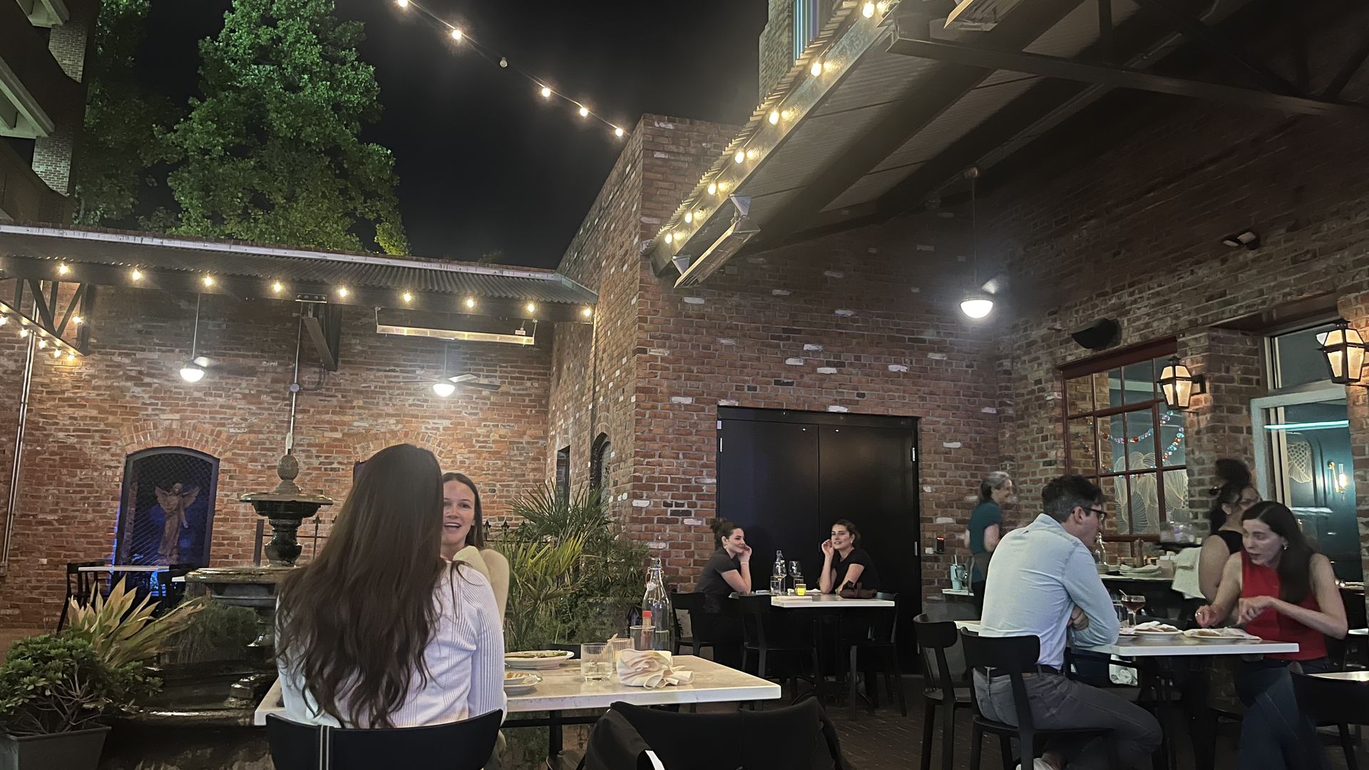 Nighttime outdoor restaurant courtyard with brick walls, string lights overhead, and groups of diners at tables. A green tree on the left and a fountain in the foreground.