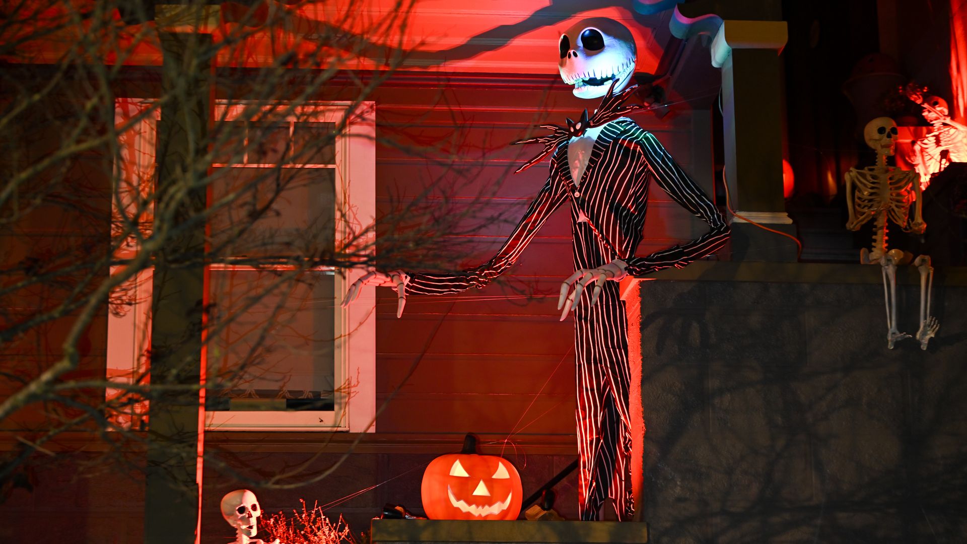 Halloween decorations at night with a large skeletal figure in a pinstripe suit, two smaller skeletons, and a glowing jack-o'-lantern, all bathed in red light on a porch.