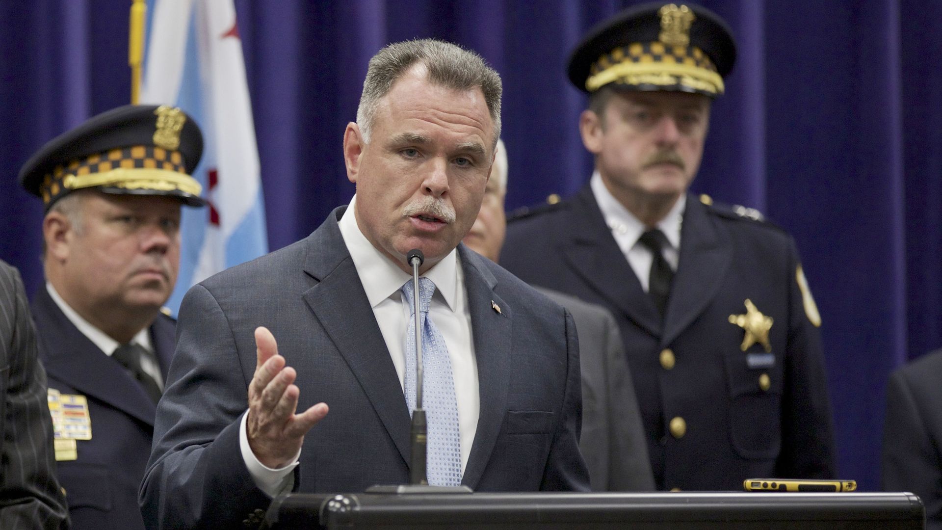 Man in a dark suit and light blue tie speaking at a podium with two police officers in uniform behind him, one with a checkerboard hat band, in front of a dark curtain.