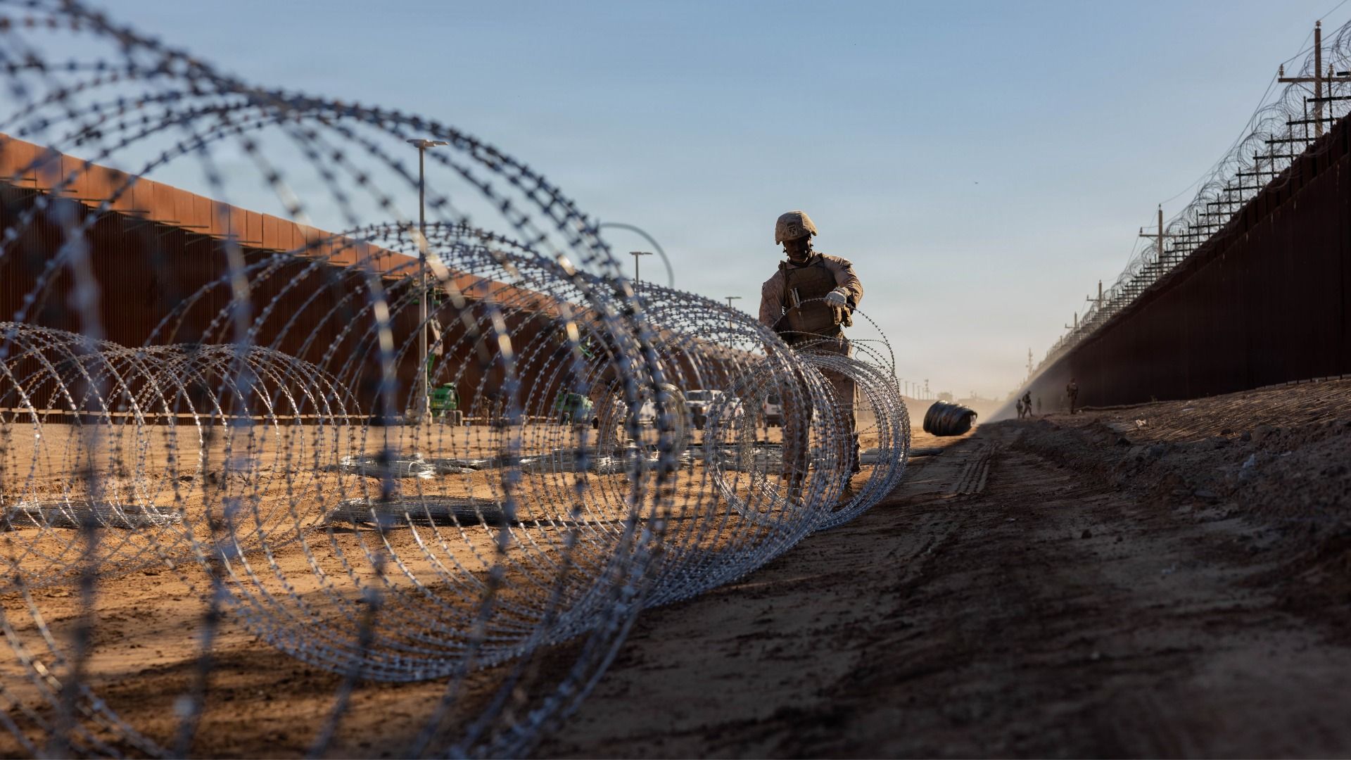 Marine in tactical gear unrolling large coils of silver concertina wire along a dirt path between two tall brown metal fences under a clear blue sky at sunset.