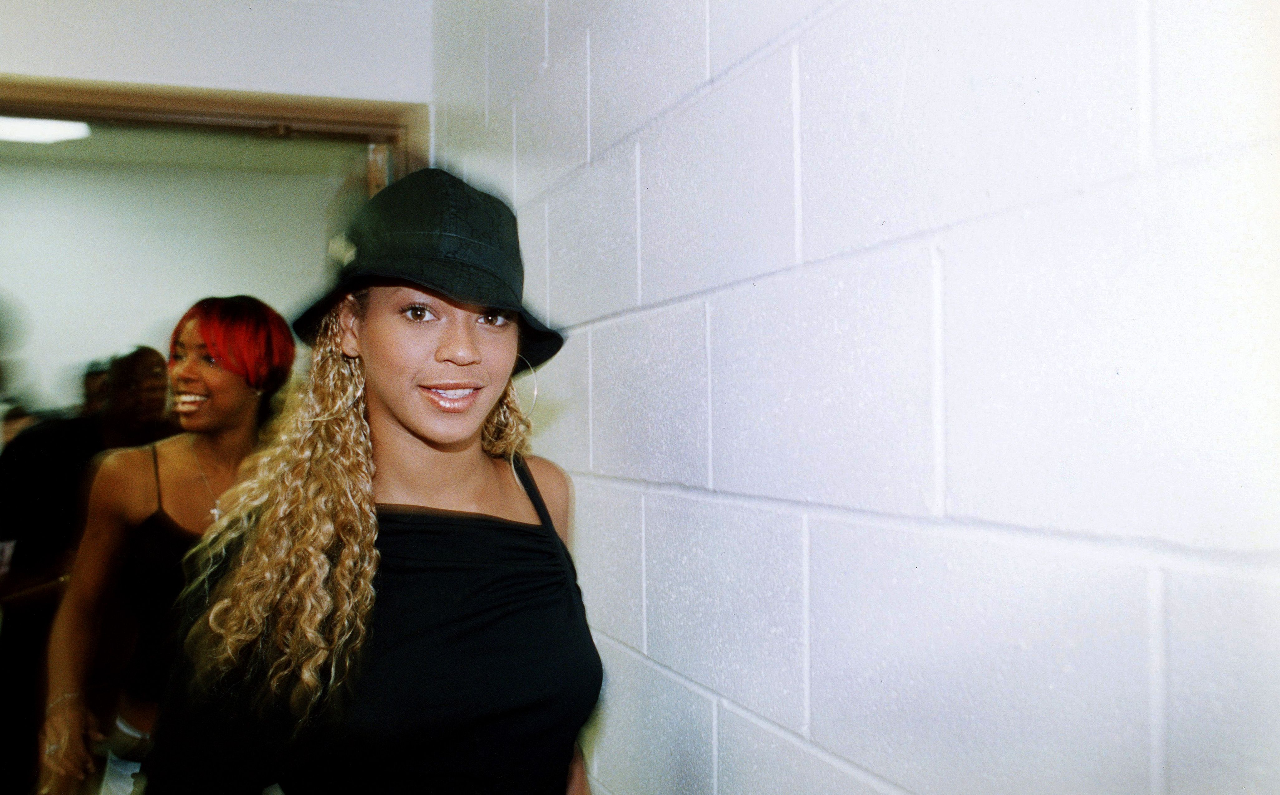 Photo of a woman walking against a white-bricked wall, smiling at camera. 