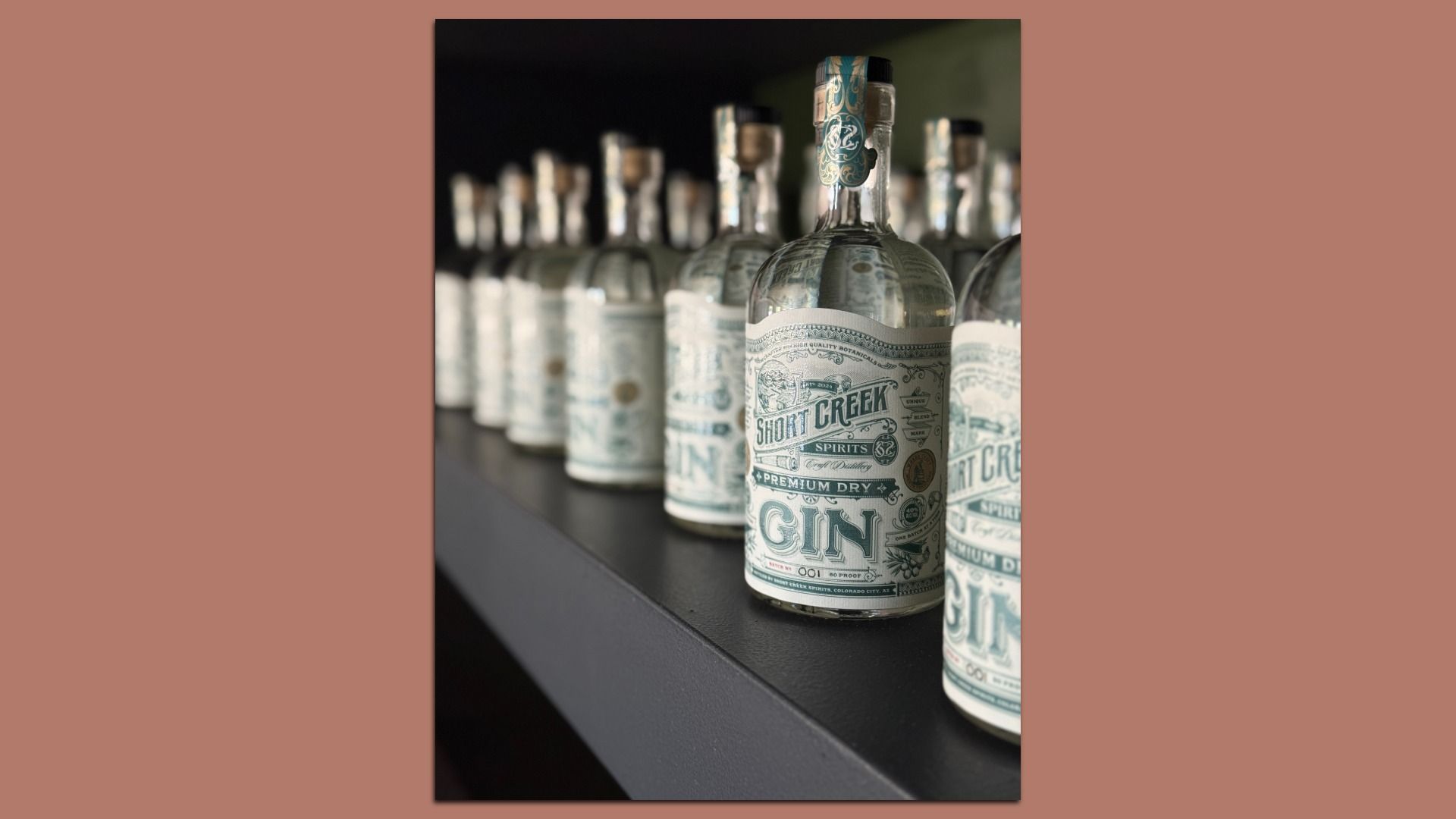 Row of clear glass gin bottles labeled "Short Creek Spirits Premium Dry Gin" with detailed blue and white labels on a black shelf, fading into background blur.