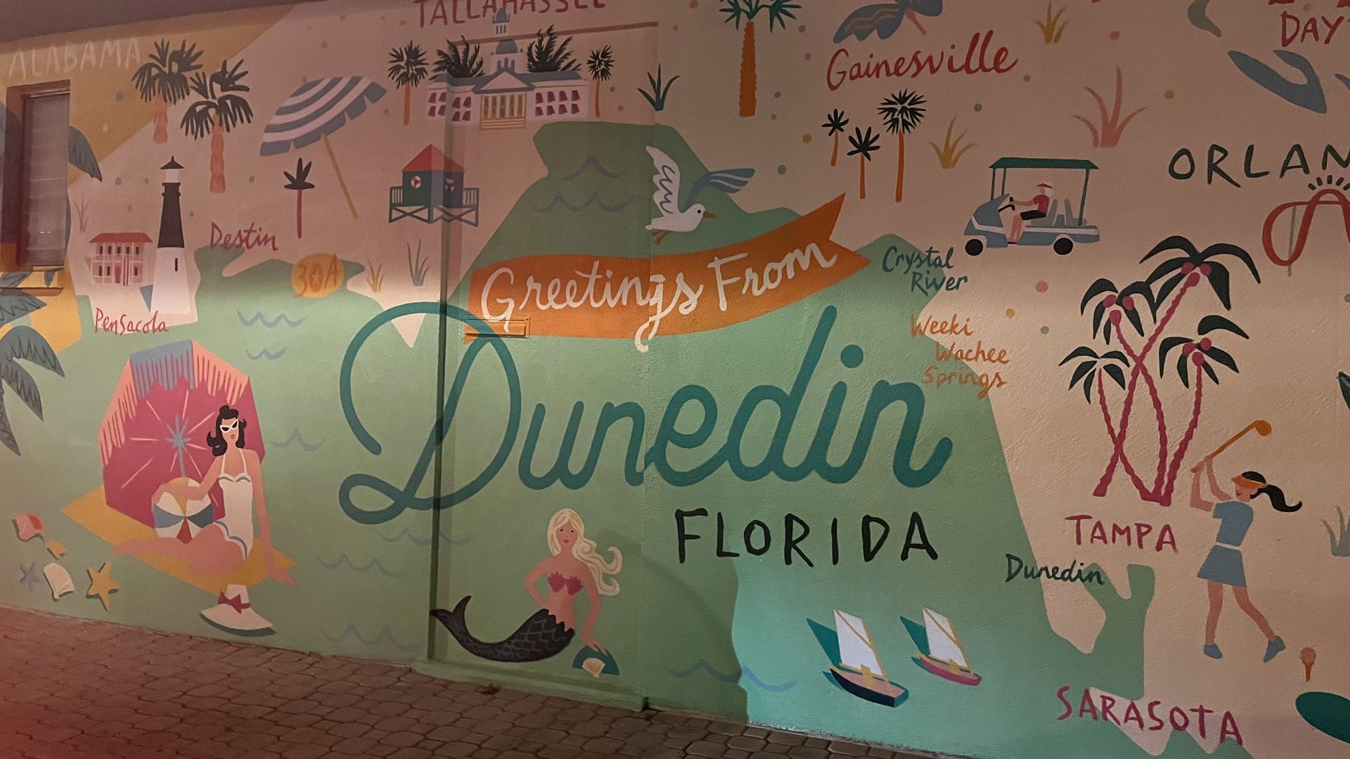 Colorful Florida wall mural with city names Orlando, Gainesville, Pensacola, Tampa, Dunedin; palm trees, boats, a mermaid, swimmers, and "Greetings From Florida" on an orange ribbon.