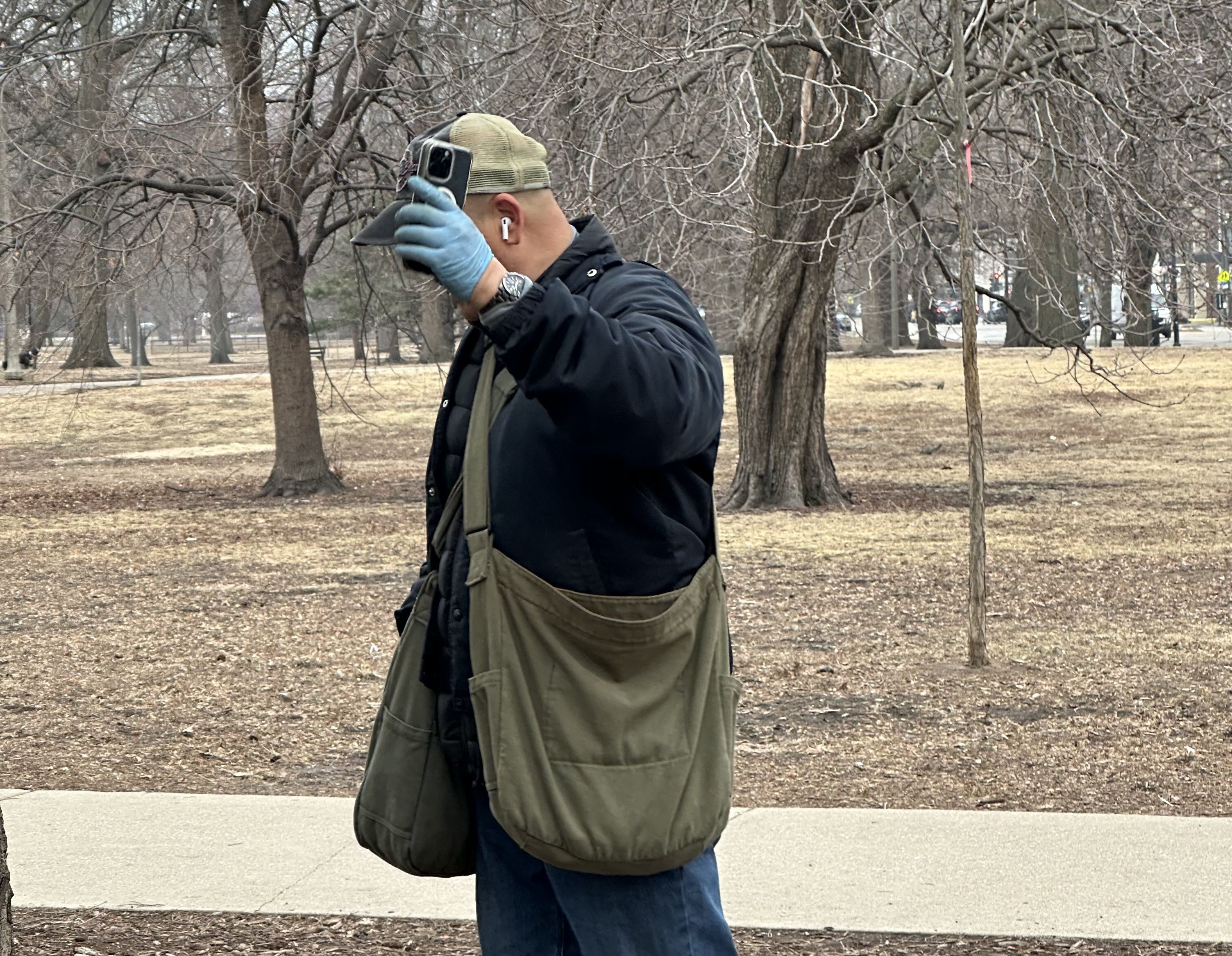 A man with cloth satchels covers his face with his phone in an outdoor park. 