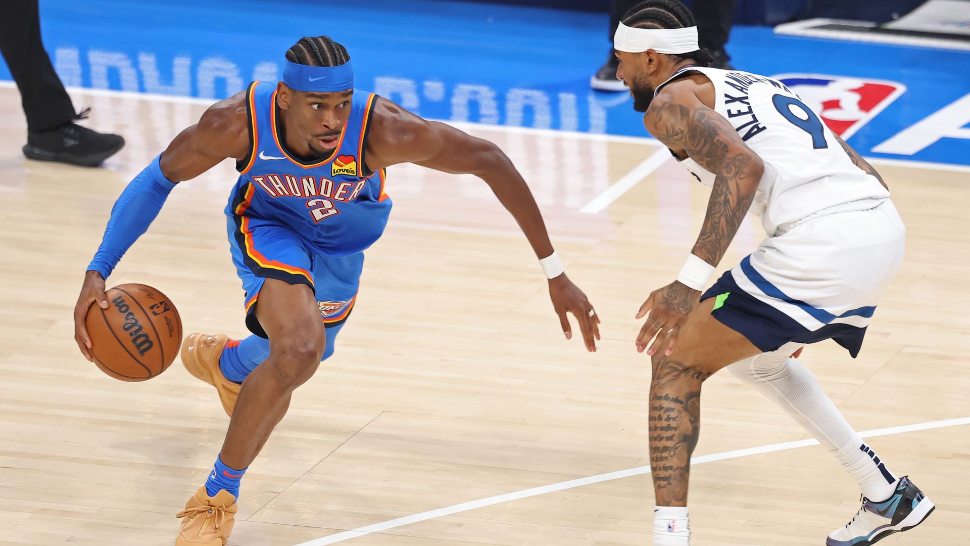 A person in a blue jersey and orange shoes dribbles a basketball around a person in a white jersey