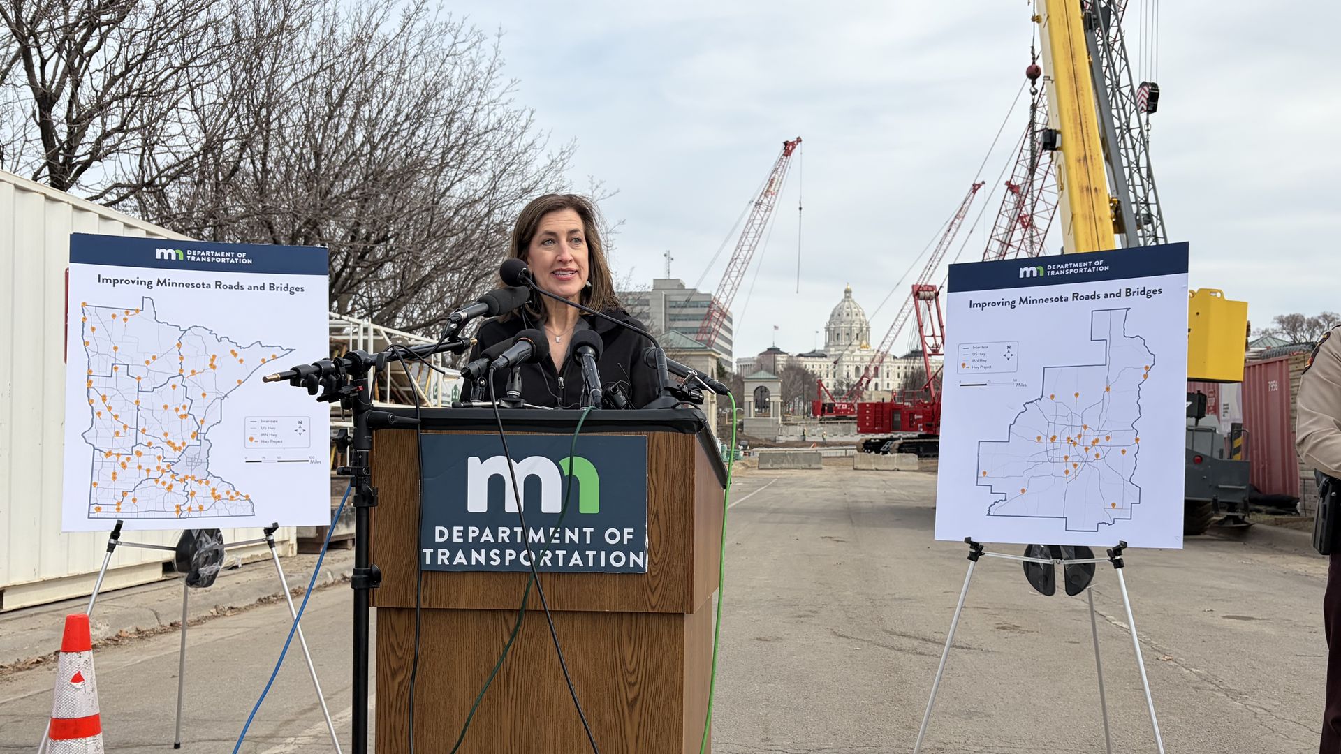 Speaker at a podium for the Minnesota Department of Transportation, with posters reading "Improving Minnesota Roads and Bridges" at a construction site with cranes and a uniformed guard nearby.