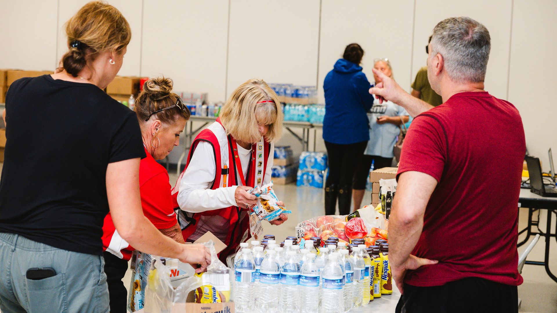 People gather around a table of snacks and bottled water in a bright room; a blonde woman in a red-and-white vest opens snack packages while others chat nearby.