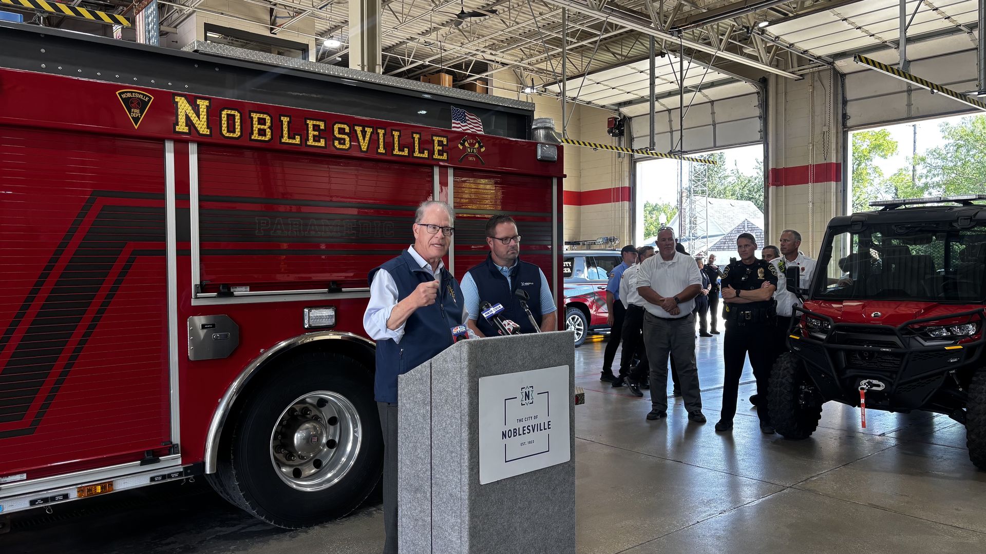 Press conference inside a fire station with a large red Noblesville fire truck featuring gold lettering; a speaker at a podium addresses officials and crew nearby.