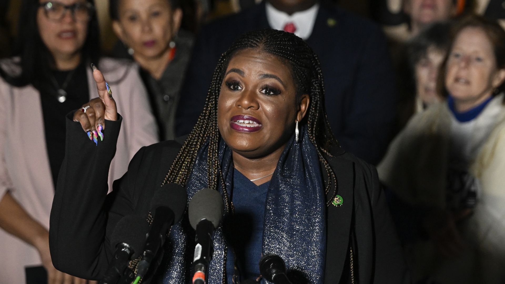 Rep. Cori Bush, wearing a black blazer, blue shirt and blue scarf, speaks in behind microphones flanked by several colleagues.