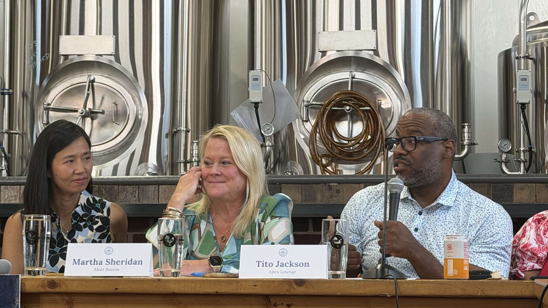 A close-up shot of three Boston leaders speaking at a roundtable at Roundhead Brewing in Hyde Park: Tito Jackson, right, holds the mic and discusses the local economy, next to Martha Sheridan, center, and Boston Mayor Michelle Wu, left.