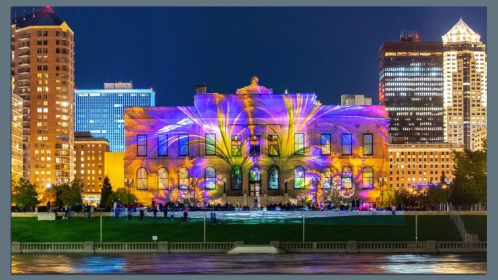 Cityscape at night with historic building lit by bright purple and yellow light projections, surrounded by illuminated modern skyscrapers and reflected in water in the foreground.