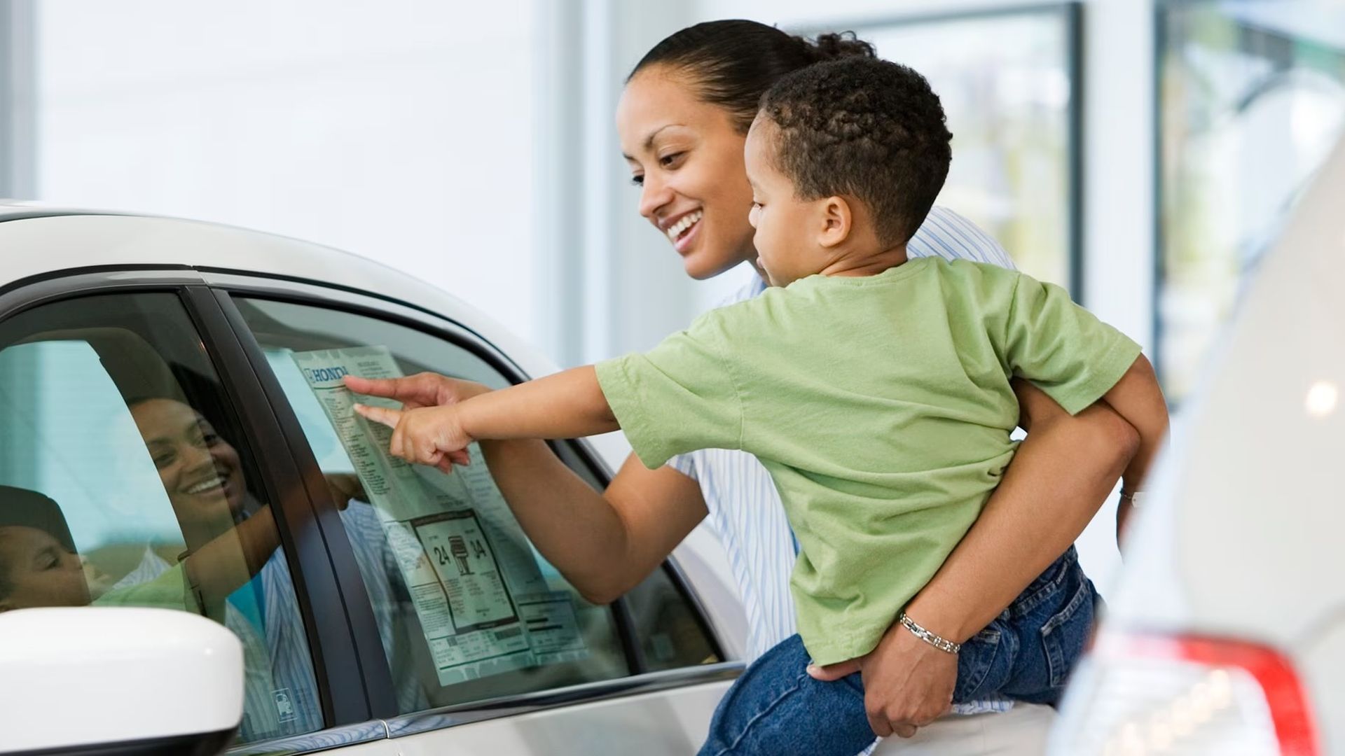 A smiling woman holds a young boy in a green shirt as they point at a car's window sticker in a bright showroom.