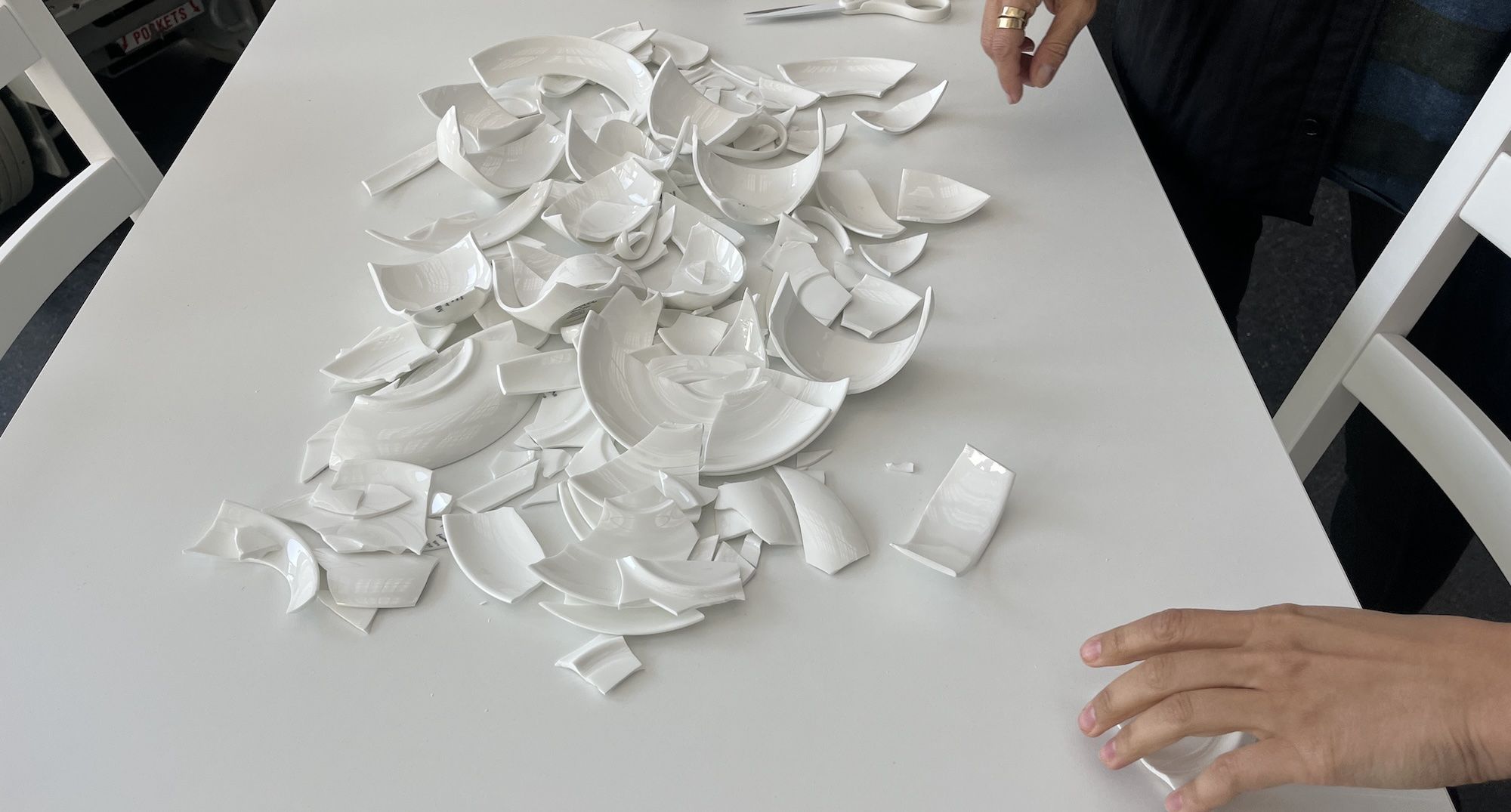 Broken white ceramic plate pieces scattered on a white table with a person's hands nearby, one wearing gold rings.