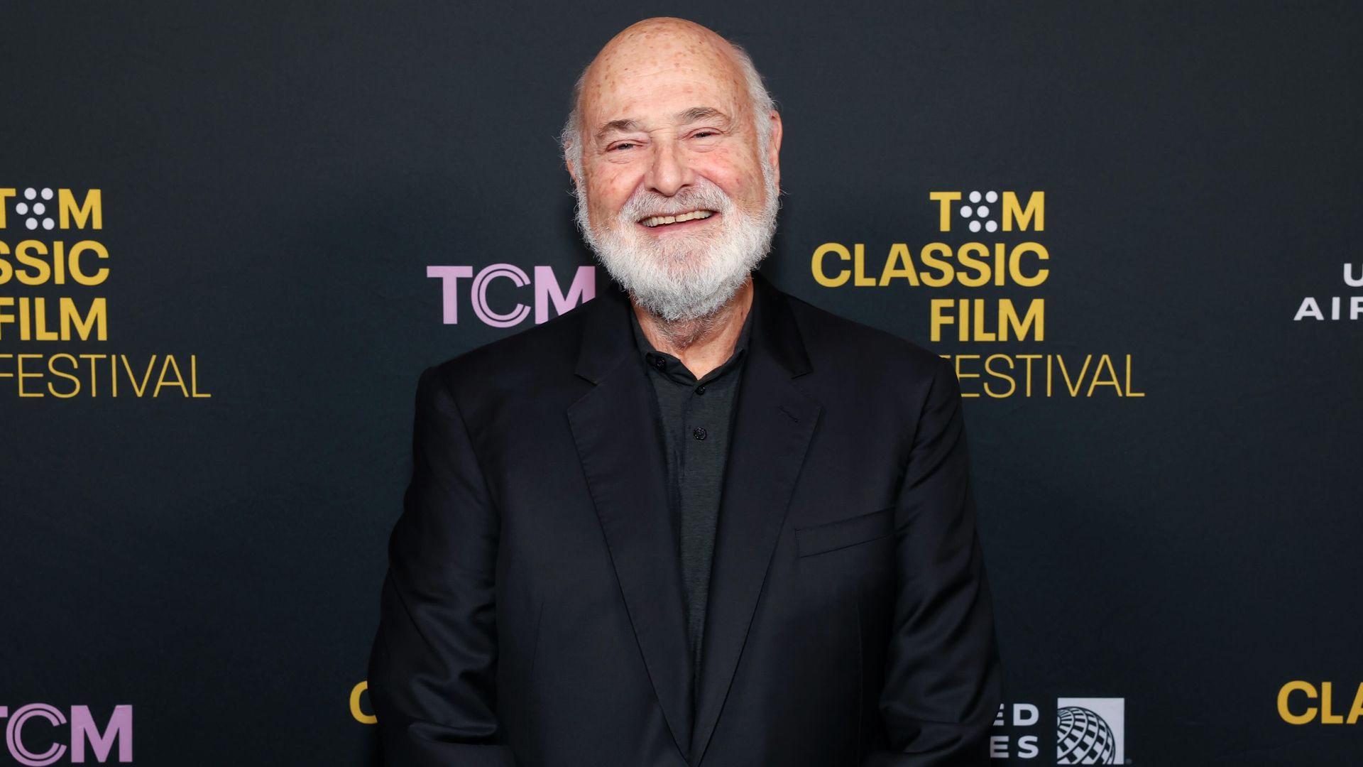 A bald, white bearded director Rob Reiner smiles while wearing a black shirt and jacket against a black backdrop with the words "TCM" in pink and "Tom Classic Film Festival" in yellow.