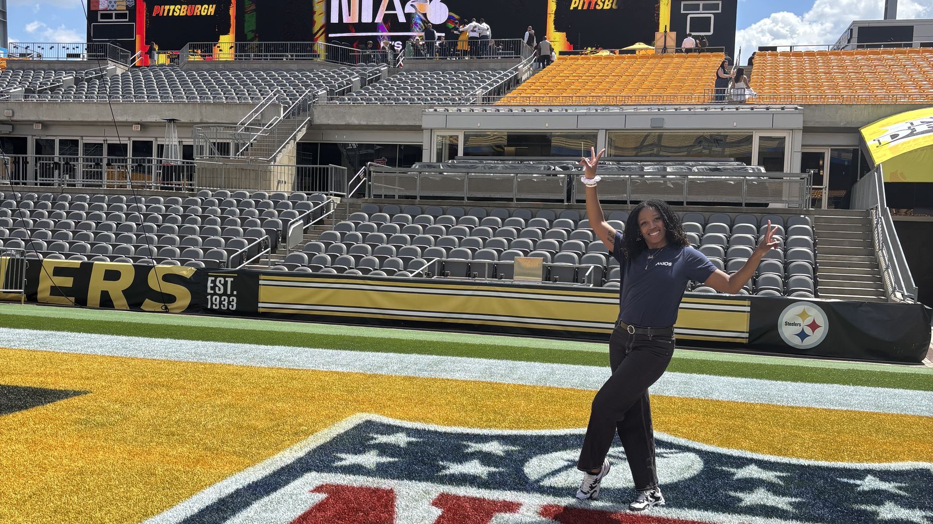 At Acrisure Stadium, a large video board shows NFL Draft graphics; a person in a blue shirt poses on the yellow end zone as empty gray and yellow seats sit in the stands under a bright blue sky.