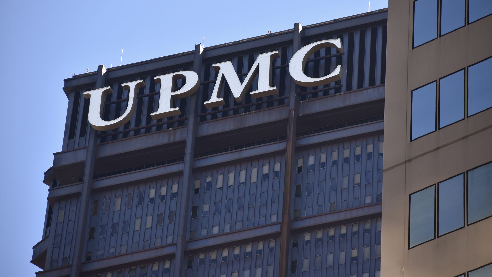 A tall modern office building with a large white sign reading "UPMC" atop the structure, blue glass windows, and a beige curved section on the right, under a clear blue sky.