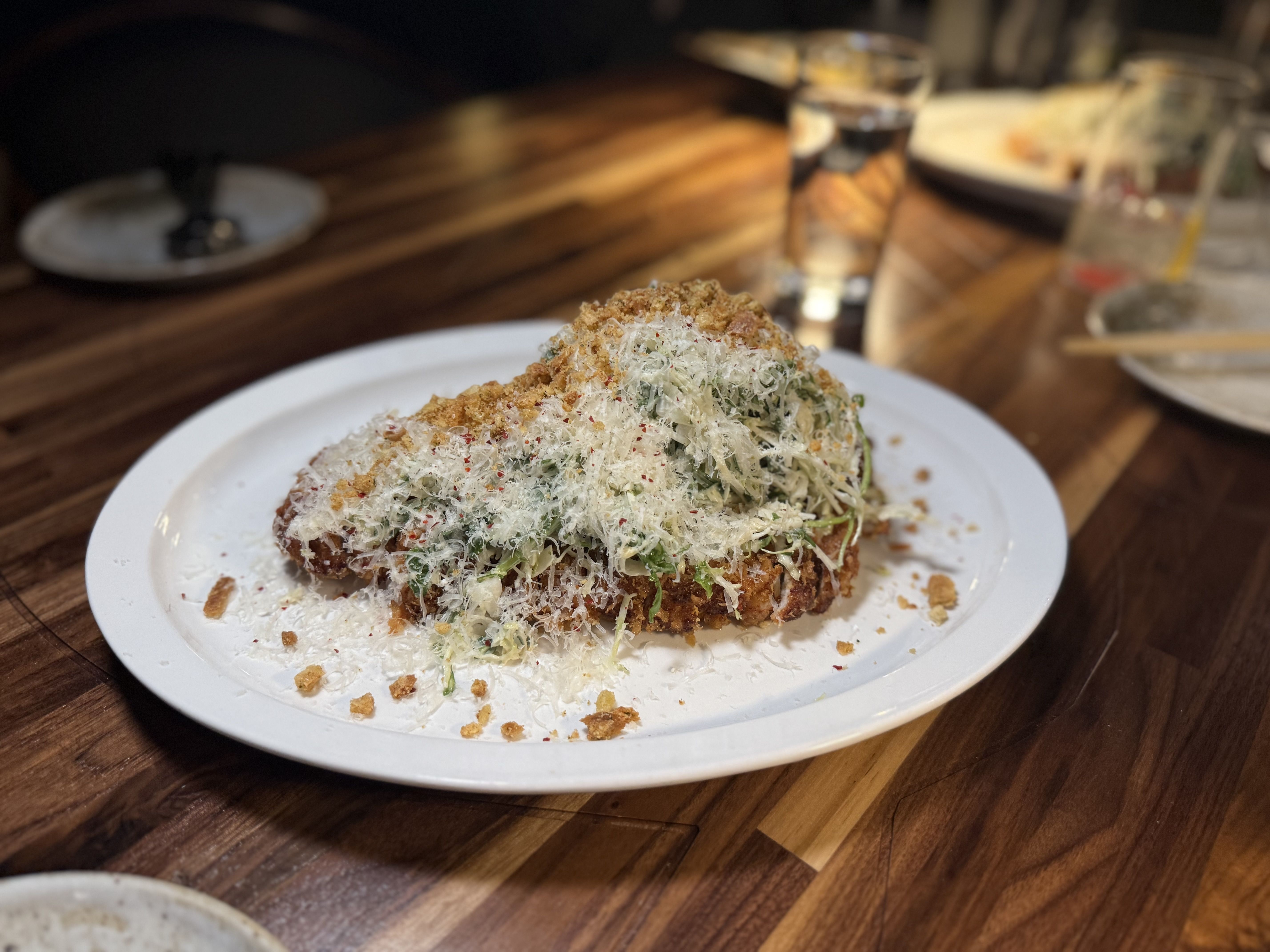 Crispy breaded dish on a white plate topped with finely shredded white cheese, green herbs, and golden brown crumbs, placed on a wooden table with blurred background elements.
