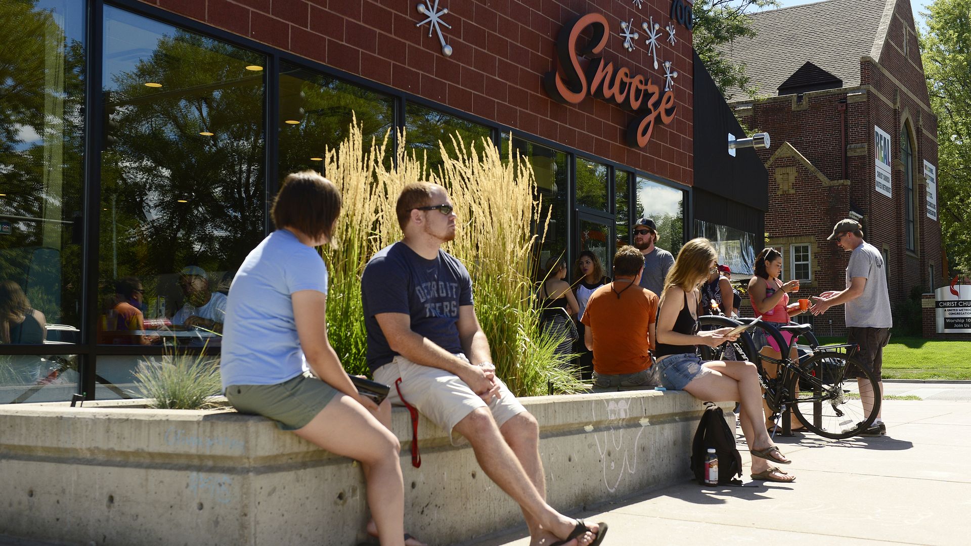 Outdoor scene of a brick storefront with a large orange "Snooze" sign and snowflake decor. People sit along a concrete planter with tall beige grasses, chatting while bikes rest nearby.