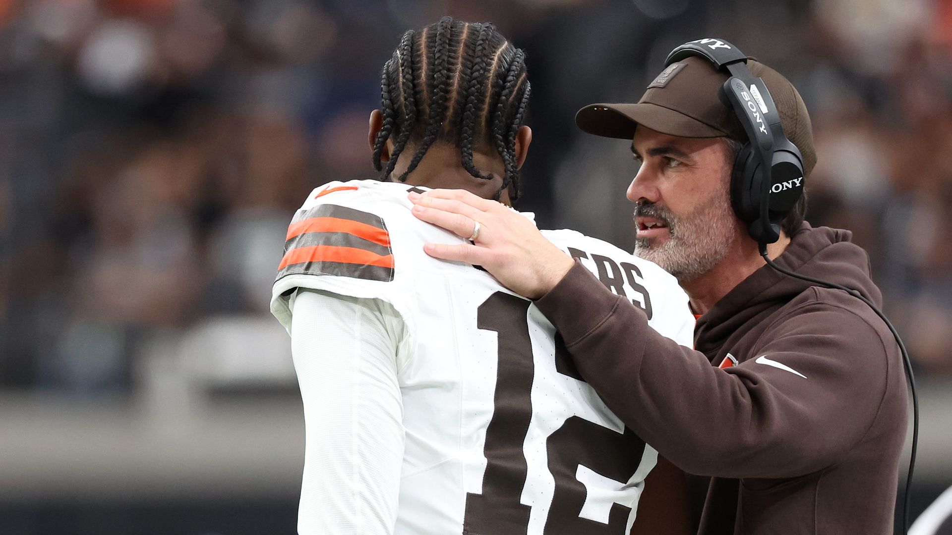 A football coach in a brown hoodie and cap talks closely to a player wearing a white jersey with number 12 and orange striped shoulder pads, placing a hand on the player's shoulder.