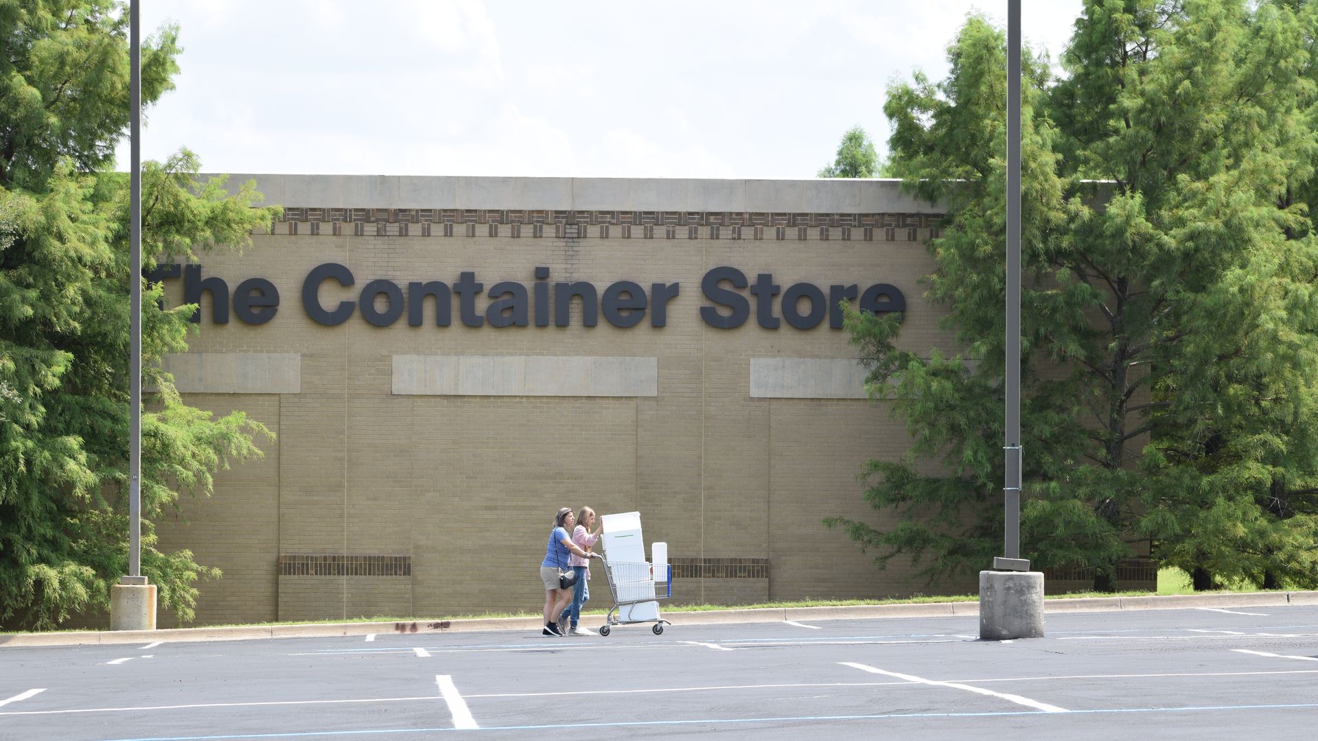 Two women push a shopping cart through an empty parking lot in front of a The Container Store location.