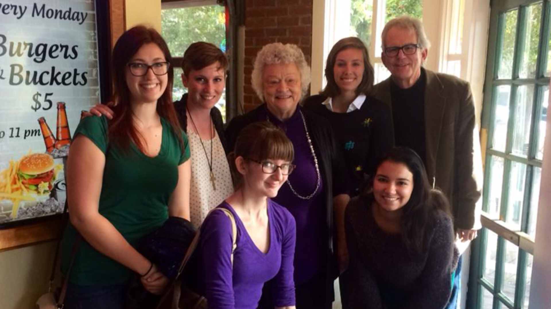 A group of seven people, six women and one man, smiling and posing for a photo at a restaurant. In the background, a poster says "Burgers and buckets $5"