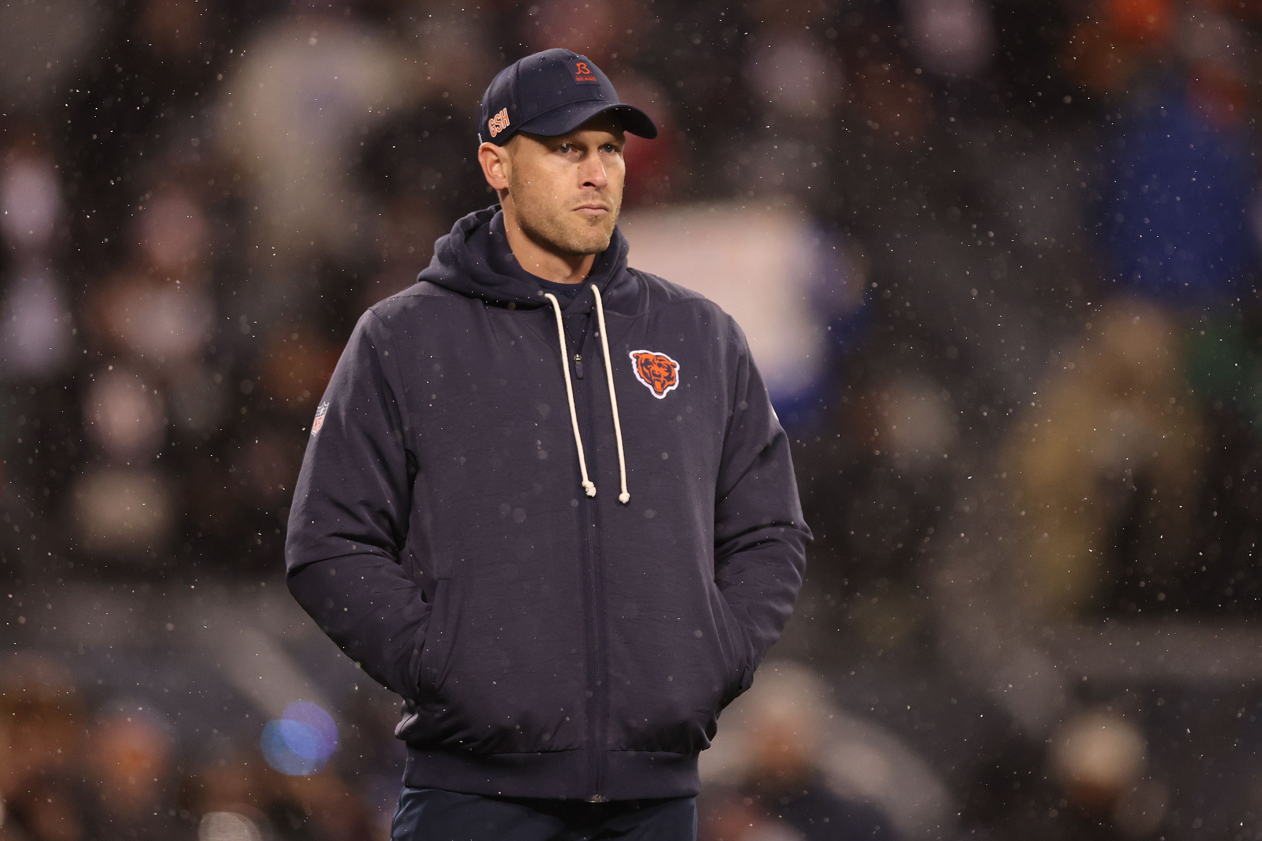 A man in a navy Chicago Bears hoodie with the team logo and cap stands outside in falling snow, looking off to the side, with a blurred crowd in the background.
