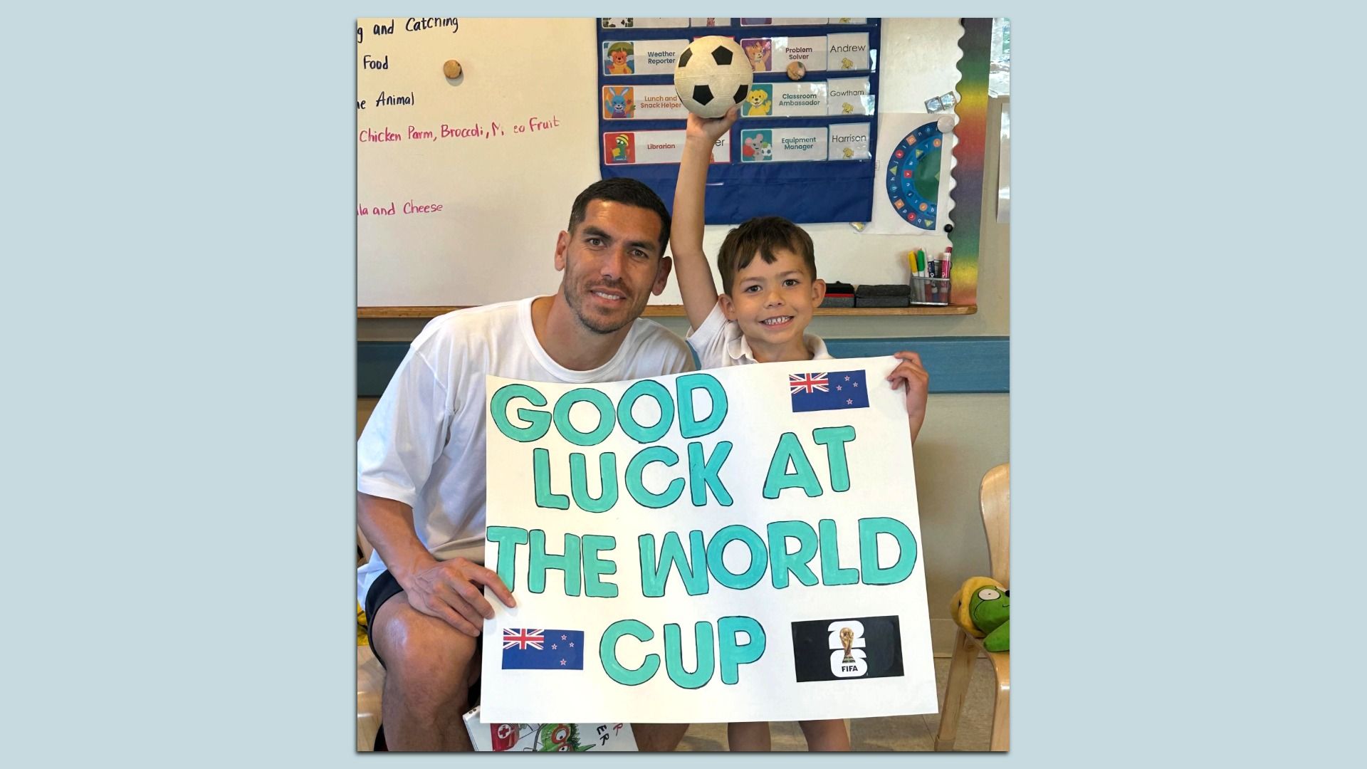 Man and boy in a classroom hold a large poster reading "GOOD LUCK AT THE WORLD CUP" with Australian flags and a FIFA logo, as the boy raises a soccer ball.