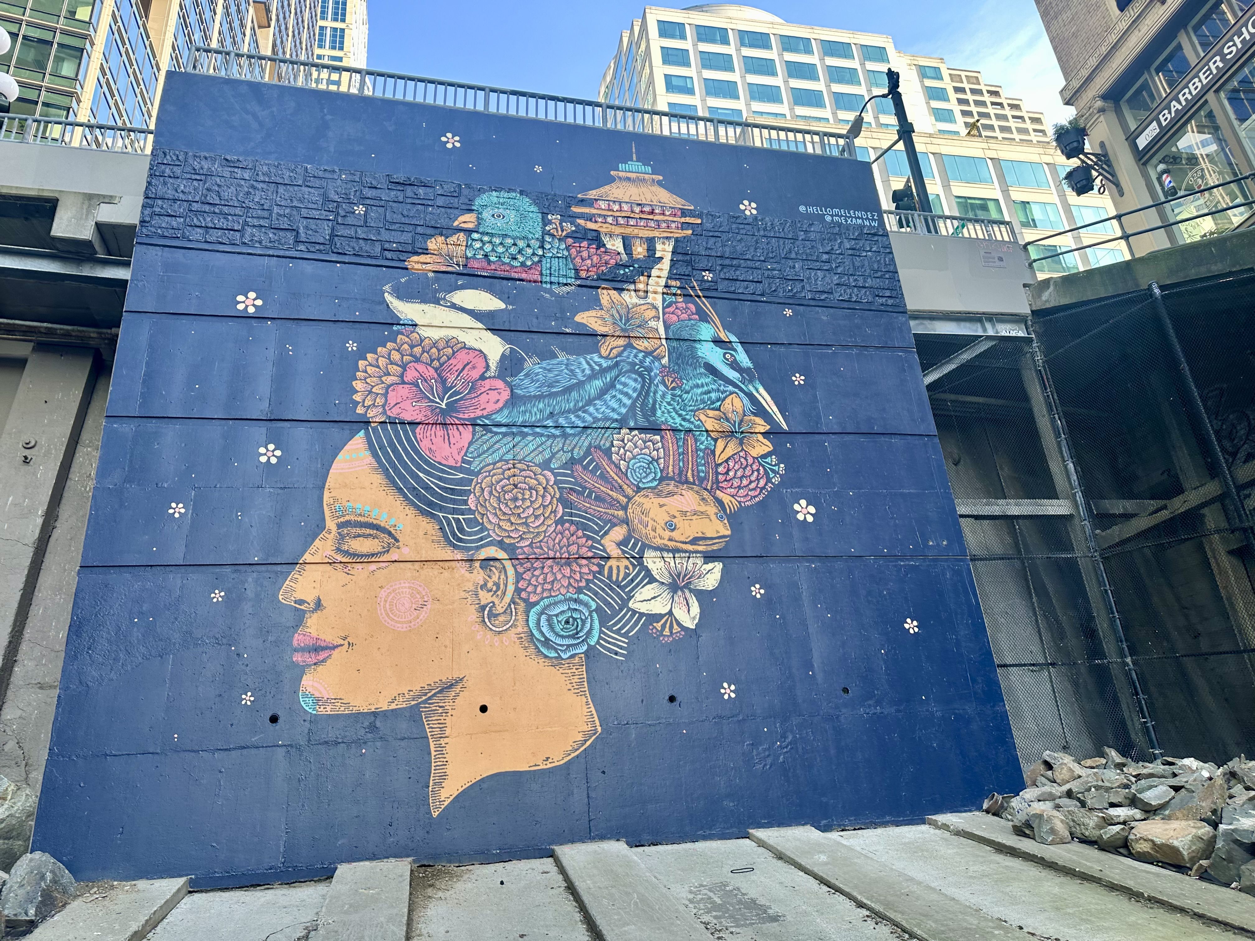 A painting of a woman with Seattle monuments and flora and fauna in her hair, with buildings on 1st Avenue visible behind the mural.
