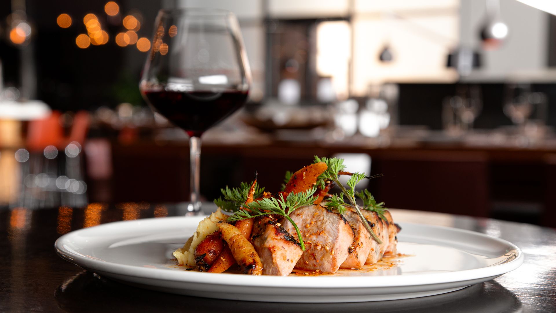 Slice of roasted meat with carrots and potatoes on a white plate, garnished with herbs, beside a glass of red wine in a dim, upscale restaurant.