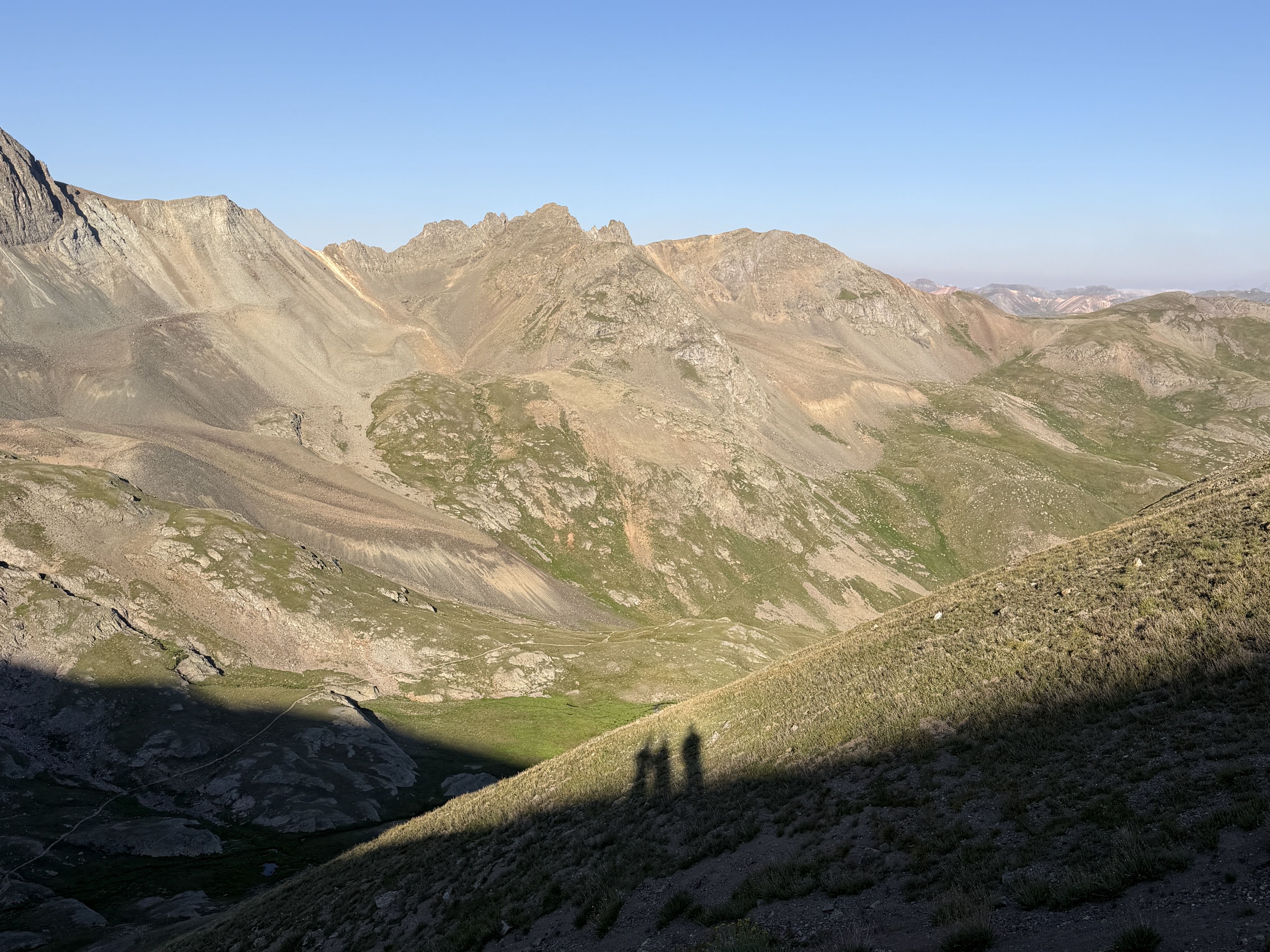 Panoramic view of jagged brown-gray mountain peaks under a clear blue sky; rocky slopes with sparse green patches, shadowy valley foreground, distant ridges on the horizon.