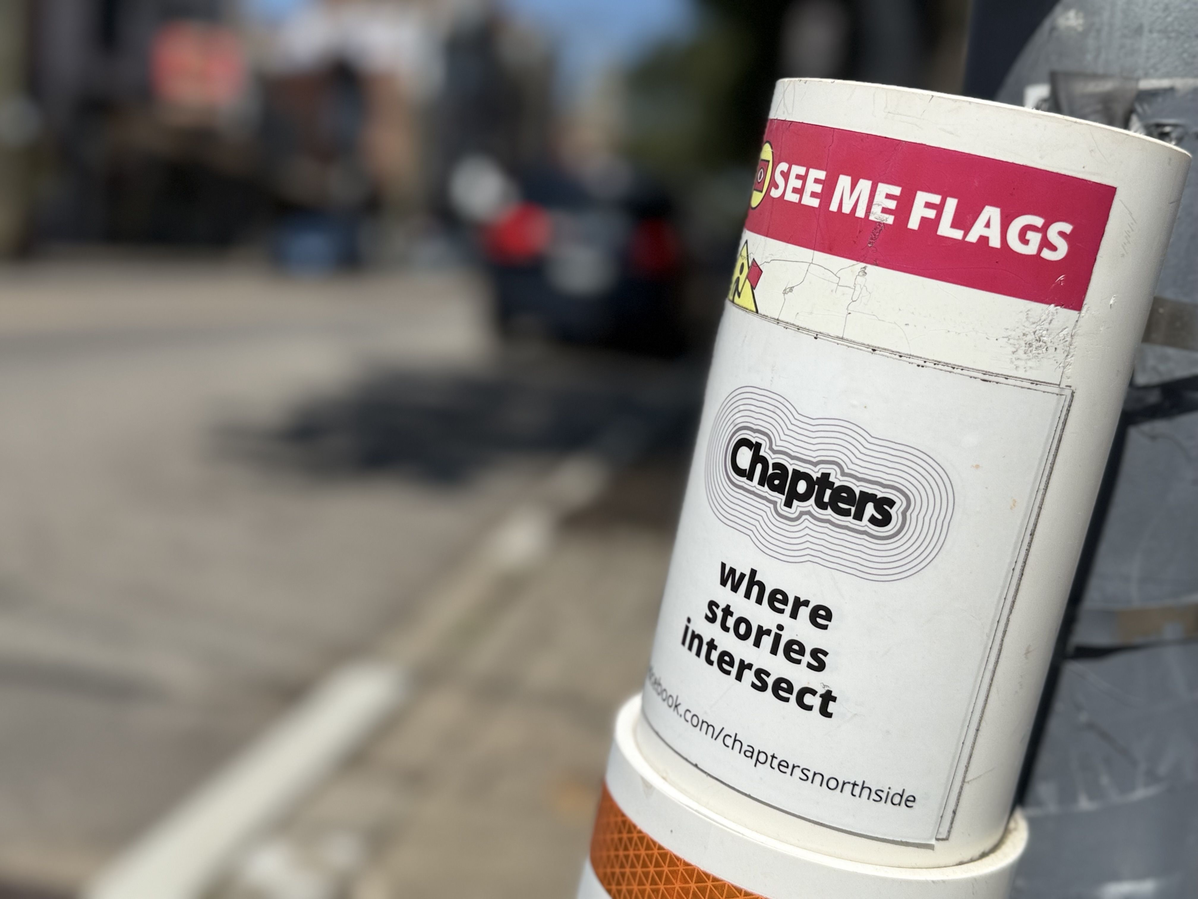 Close-up of a white traffic post wrapped with a pink banner reading "SEE ME FLAGS", with a smaller "Chapters" sticker below; a blurred urban street scene in the background.