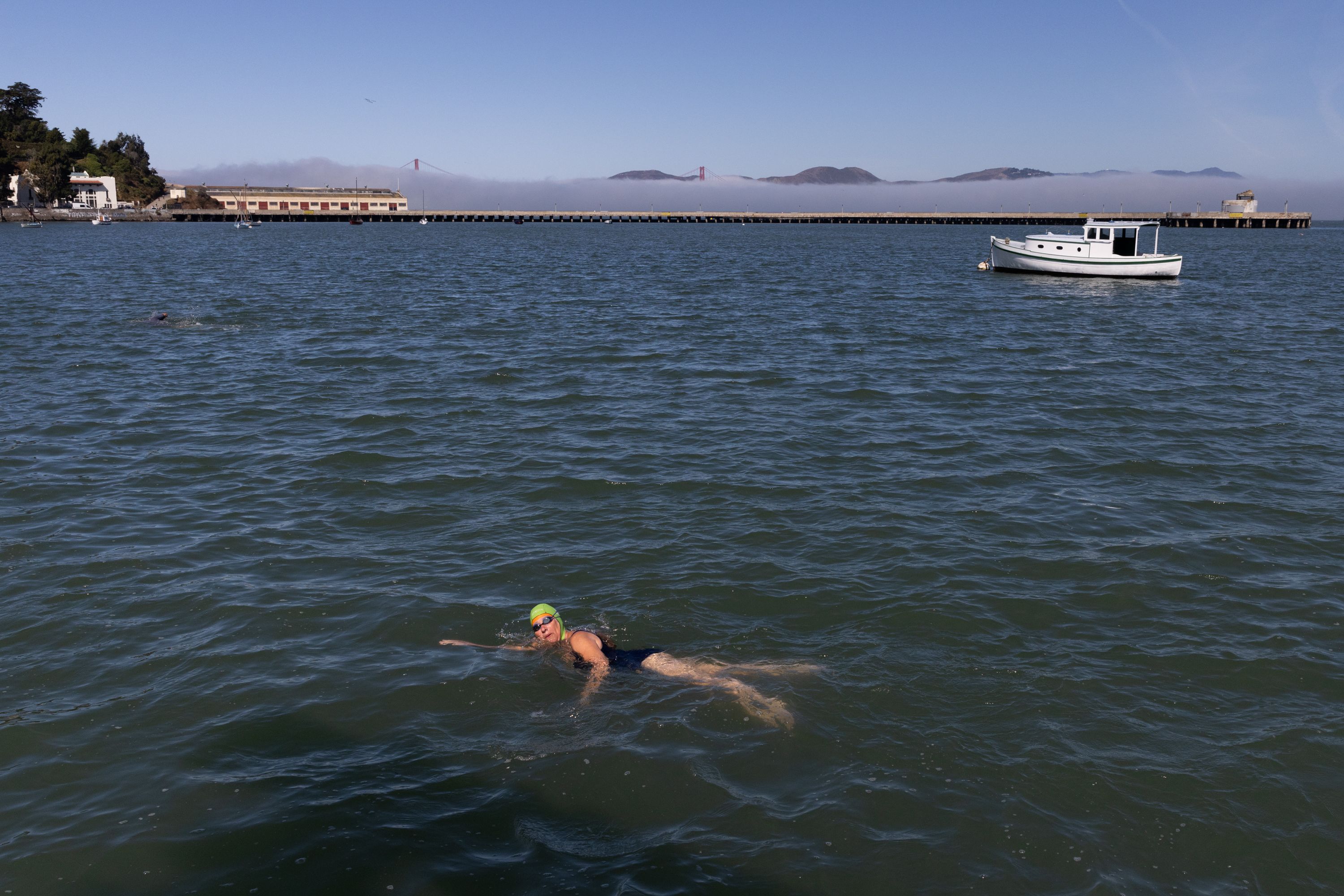 Swimmer in green cap and black swimsuit swimming in open water with a boat nearby, a pier, and fog partially covering the Golden Gate Bridge under a clear blue sky.
