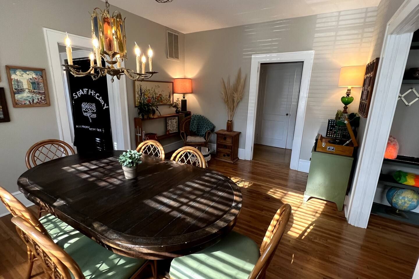 Dining room with dark wooden oval table, six wicker chairs with green cushions, sunlight casting shadows, two lamps, a chair with a blue blanket, wall art, and wooden floor.