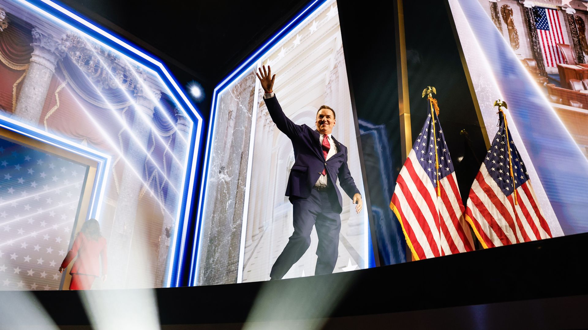 A man in a dark suit and red tie waves from a stage, flanked by American flags. Large display screens behind him show government imagery with blue neon lighting.