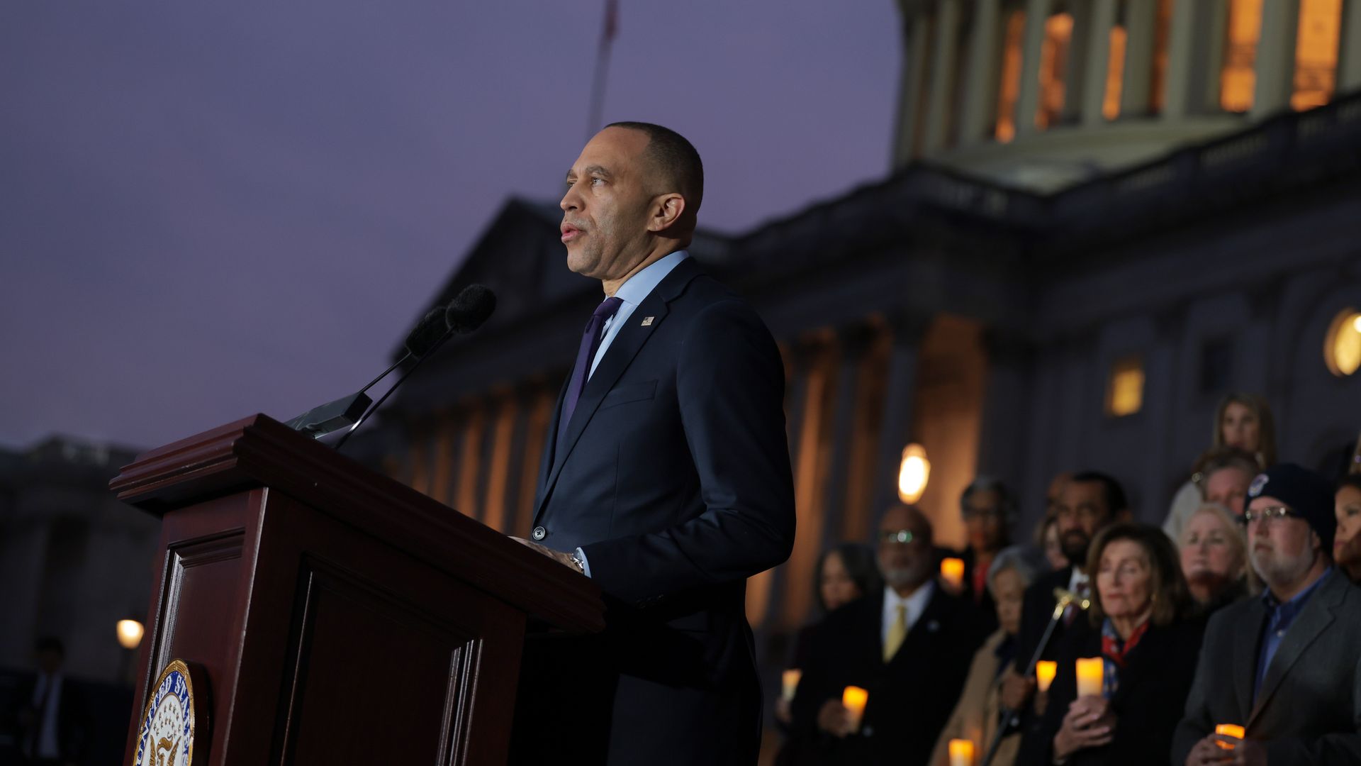 Man in a dark suit speaking at a podium with U.S. Capitol building illuminated behind him at dusk, while a crowd holding candles listens attentively.