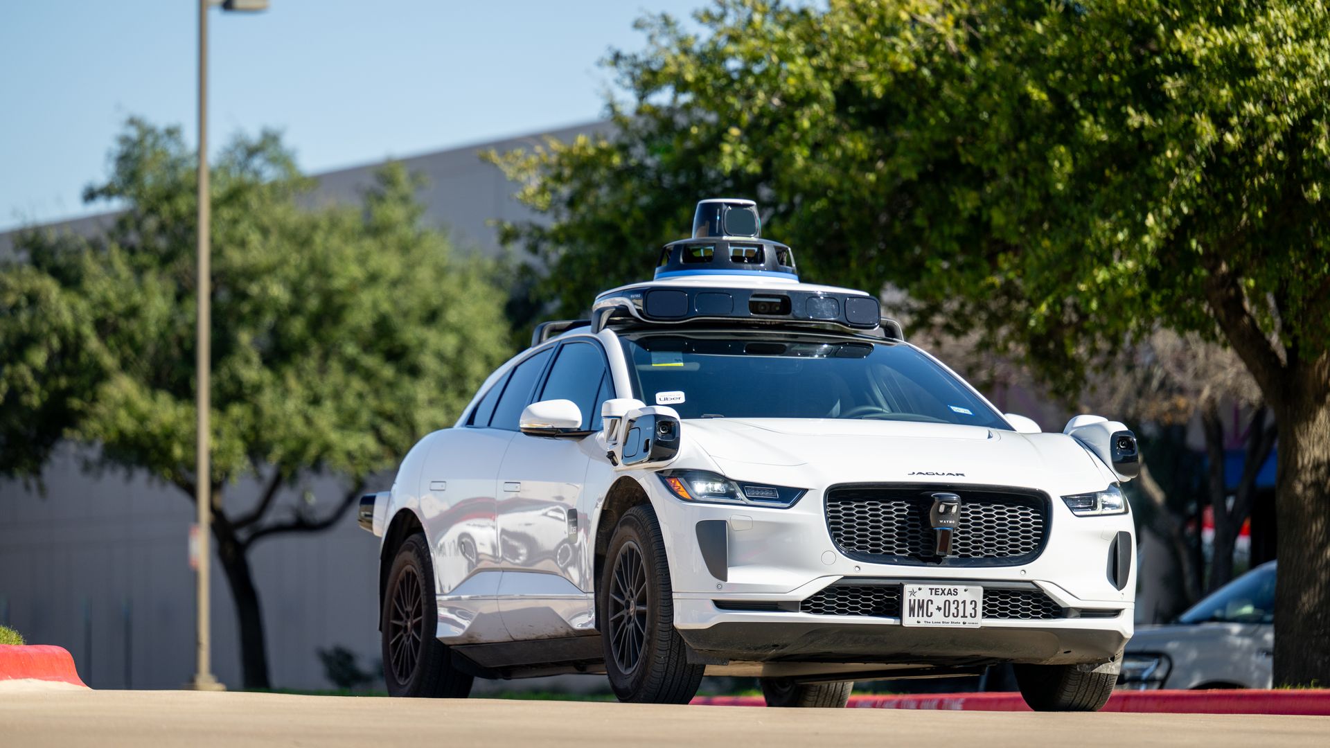 White Jaguar electric car equipped with autonomous driving sensors, driving on a road with trees and a lamppost in the background, Texas license plate visible.