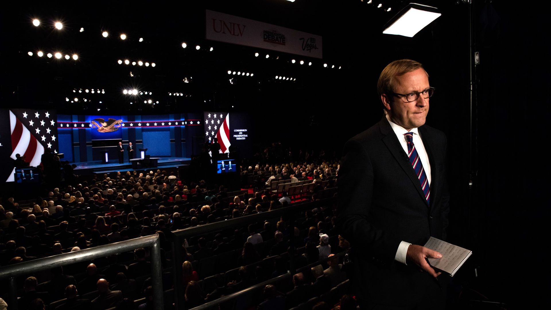In this image, Jonathan Karl stands and holds a book with a debate stage and crowd behind him.