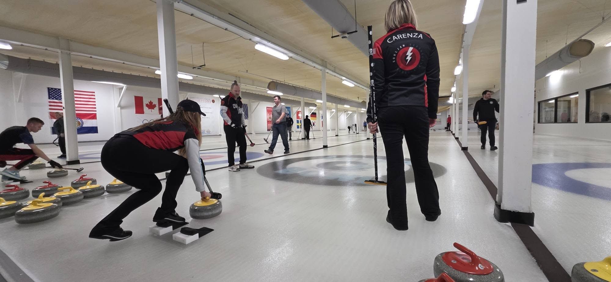 Indoor curling rink with players in black and red jackets preparing to throw stones on ice marked with circles. Flags hang on the wall; people stand and watch in the background.