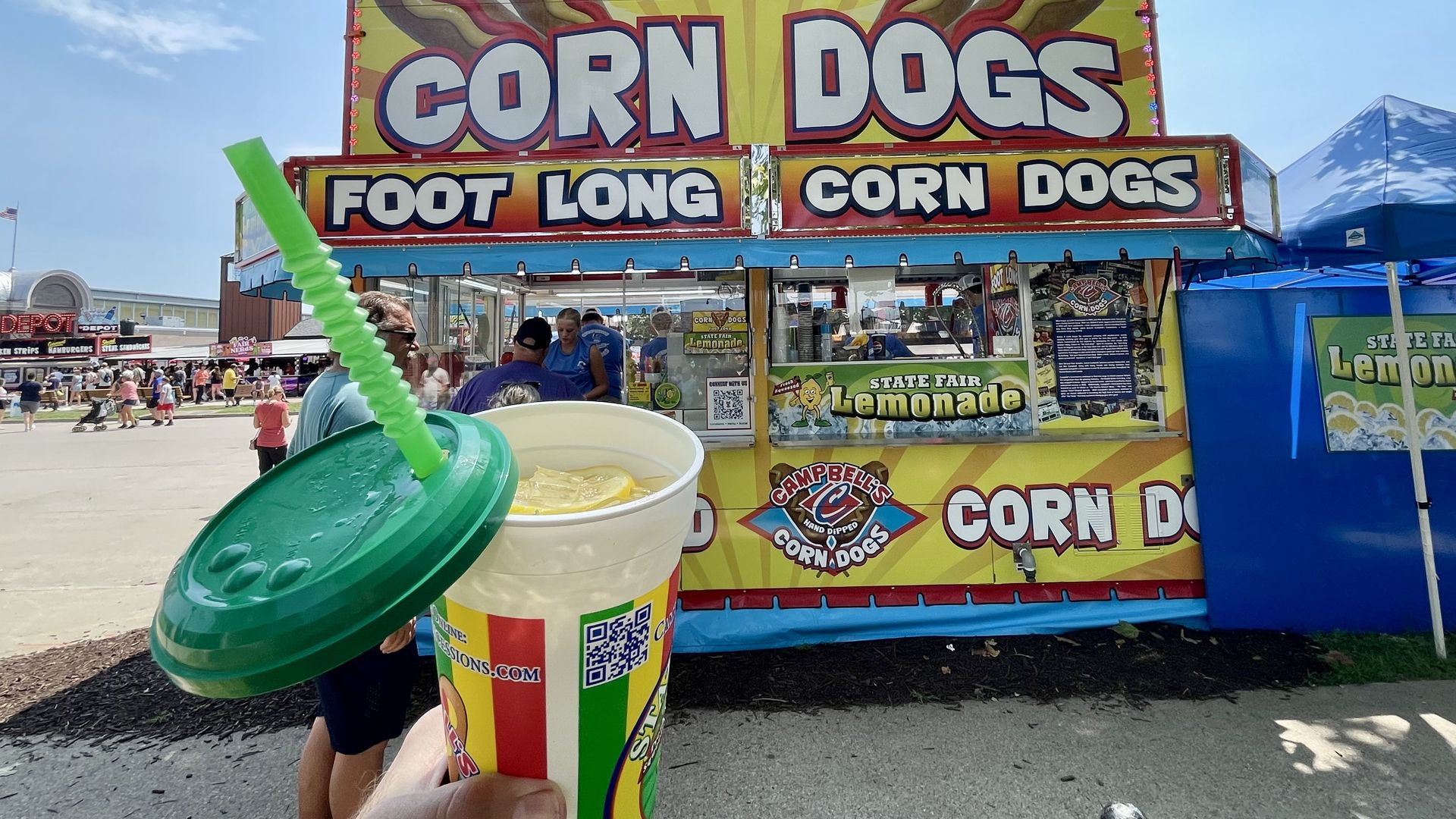 Hand holding a cup of lemonade with lemon slices and a green lid and straw in front of a colorful corn dogs food stand at a busy fair on a sunny day.