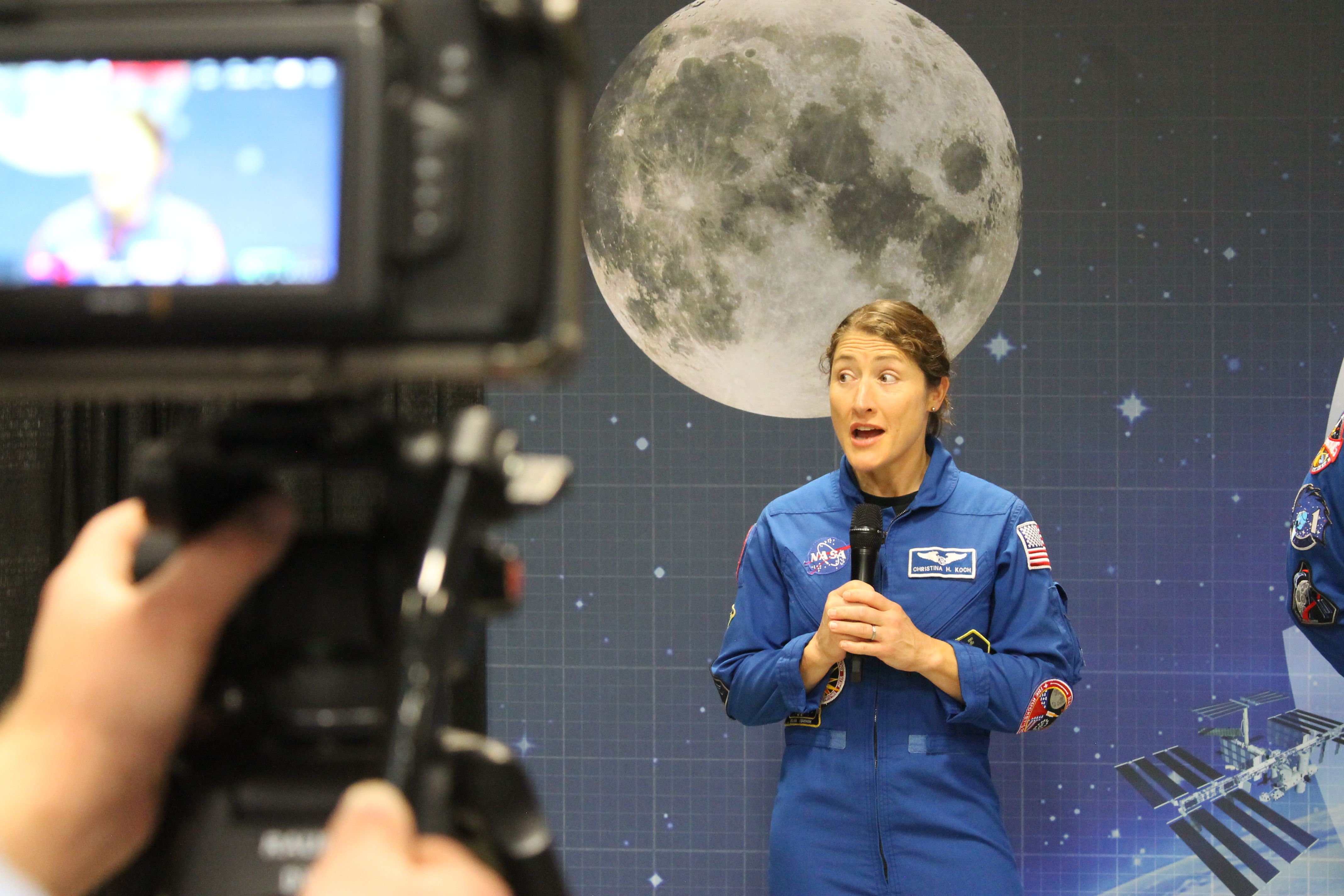 NASA astronaut Christina Koch in a blue jumpsuit speaks into a microphone during a presentation, standing in front of a large moon mural with a blurred camera in the foreground.