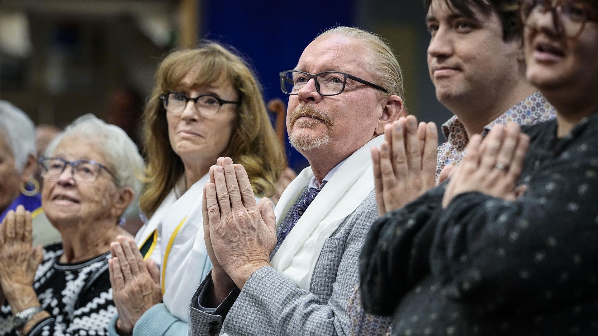 A group of people sitting in a row, holding hands pressed together as if in prayer, wearing glasses and various clothing including patterned sweaters and a gray jacket.