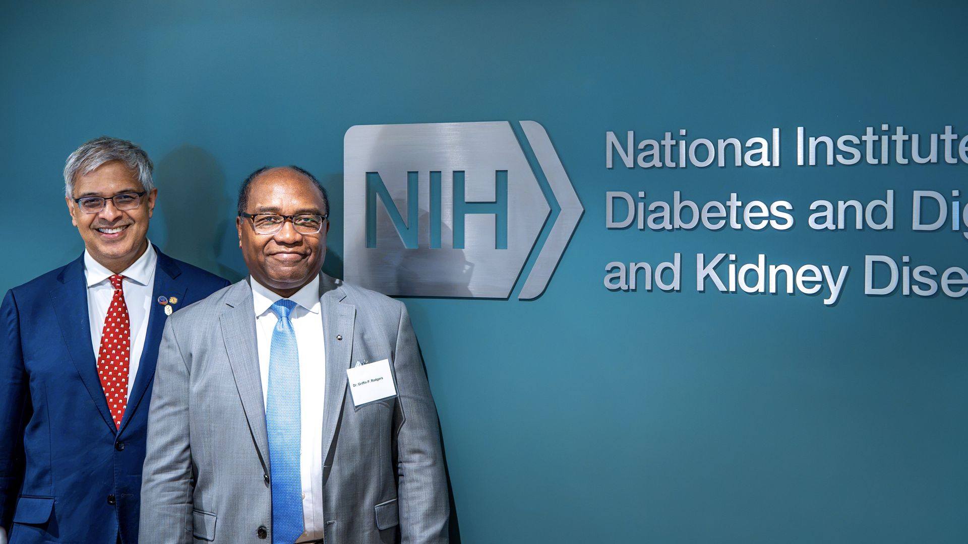 Two men in suits stand smiling in front of a teal wall with the NIH logo and the sign National Institute Of Diabetes and Digestive and Kidney Diseases.