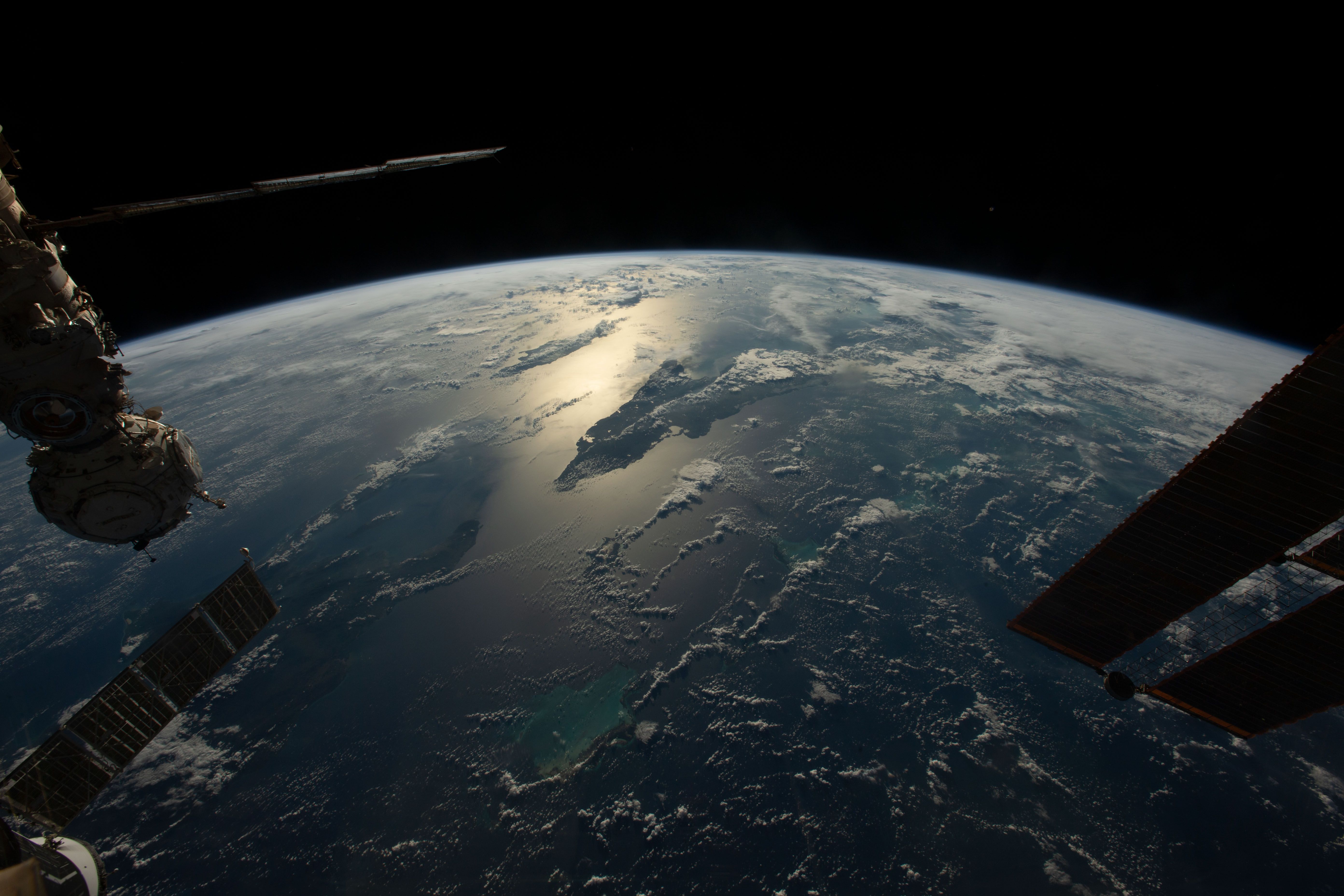 View from orbit: Earth's curved horizon, blue atmosphere, and sunlit ocean with clouds. Foreground shows parts of the space station and solar panels extending into the frame.